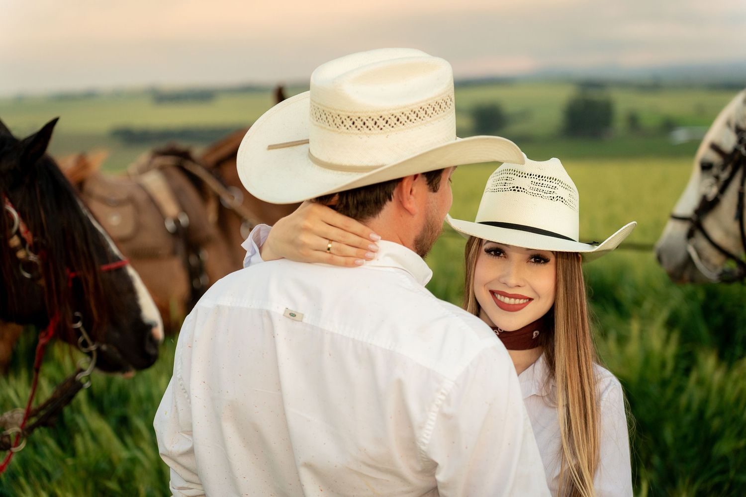 Couple in white shirts and cowboy hats posing near horses in a green field at sunset.