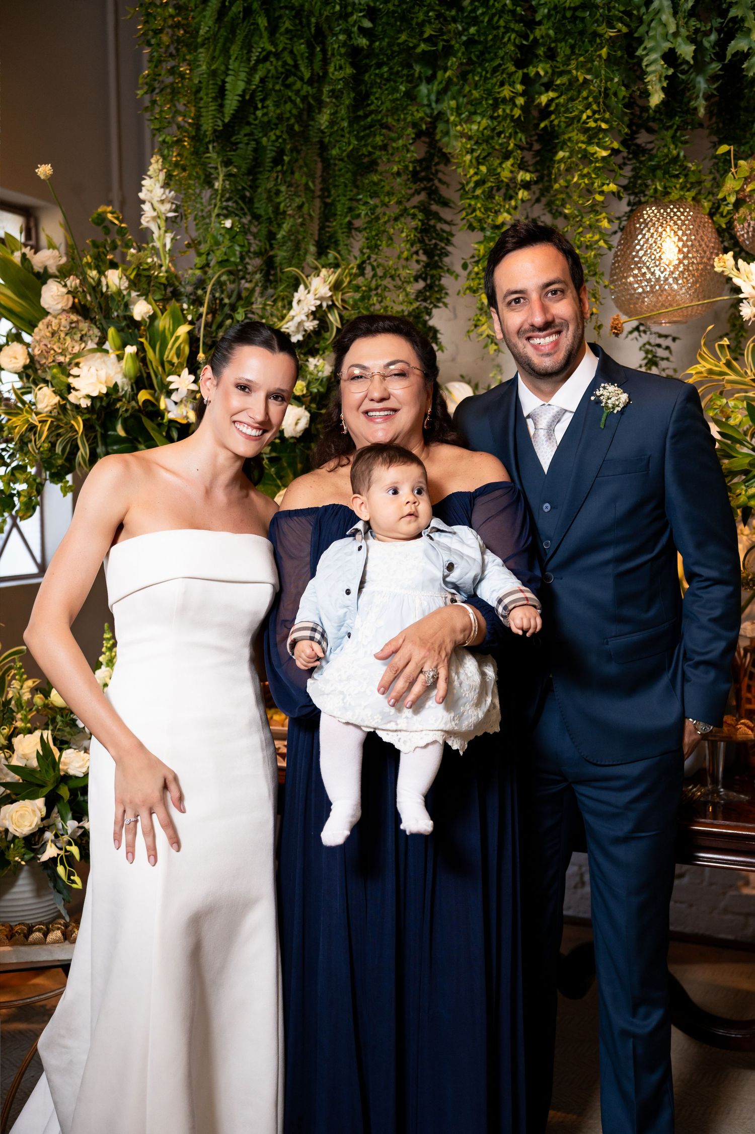Wedding party poses with baby, bride in white gown, groom in navy suit, and woman in blue dress, surrounded by lush floral decor.