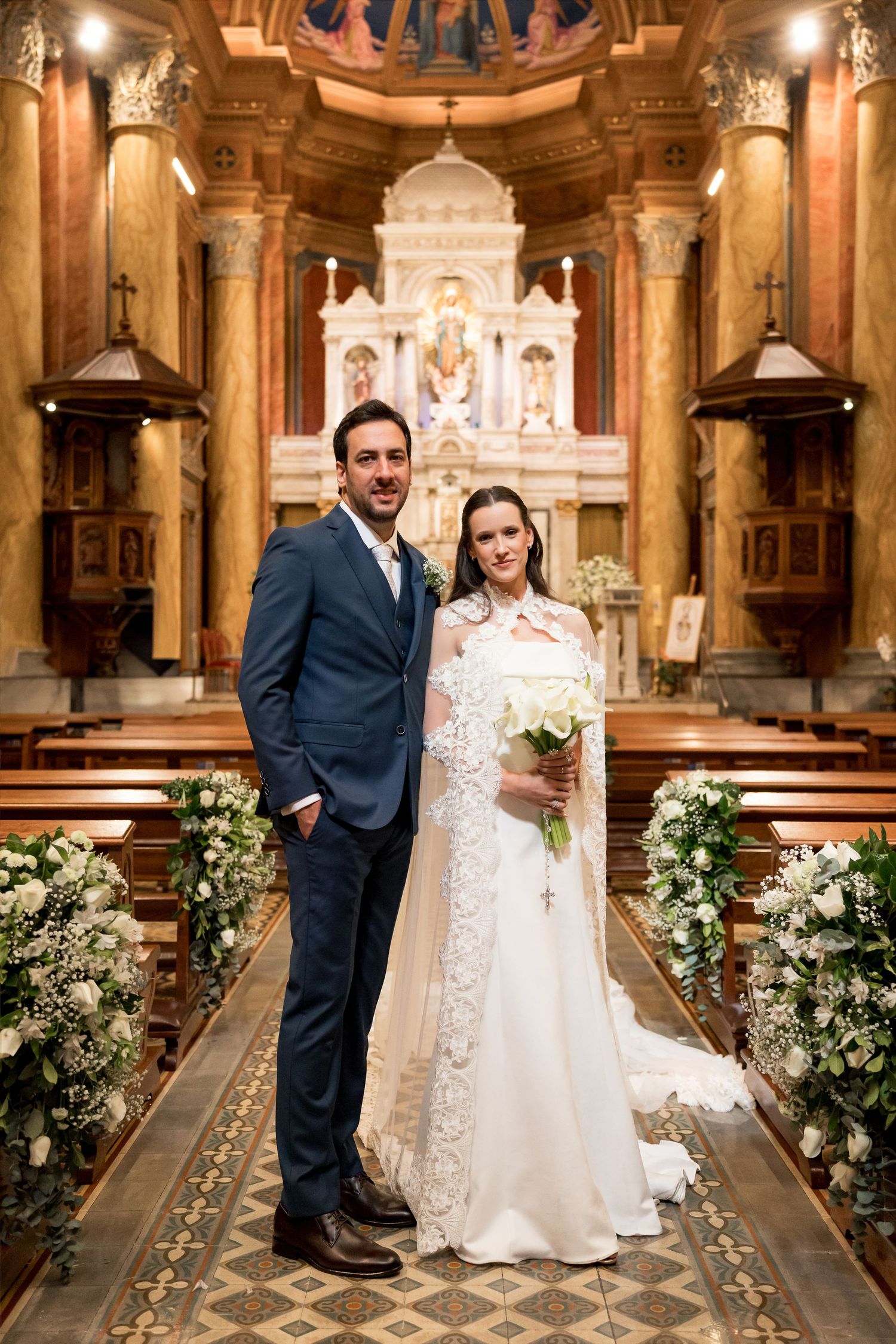 Bride and groom posing together in an ornate church aisle adorned with white floral arrangements on their wedding day.
