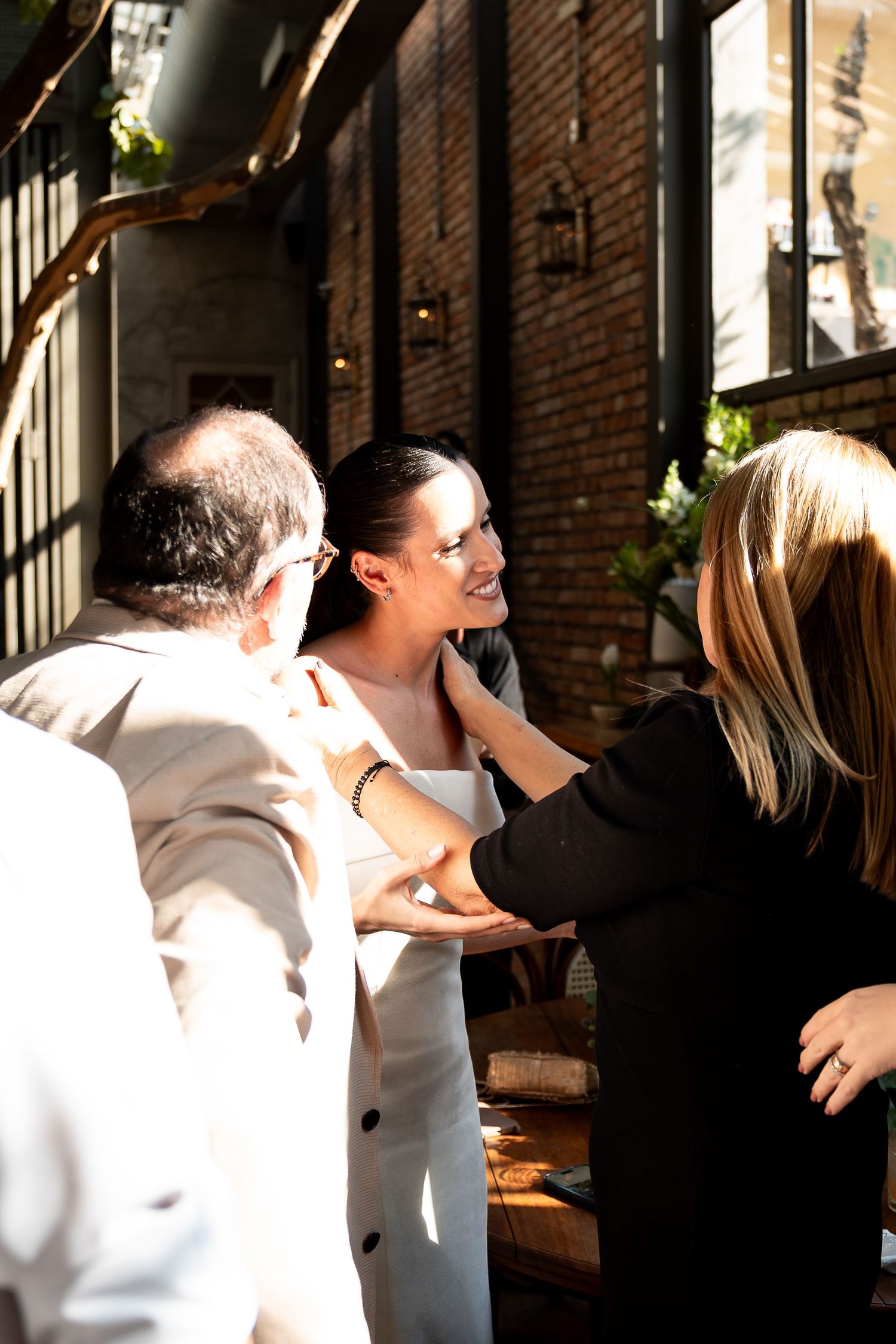 A smiling bride in white greets guests outdoors at a sunny brick-walled venue.