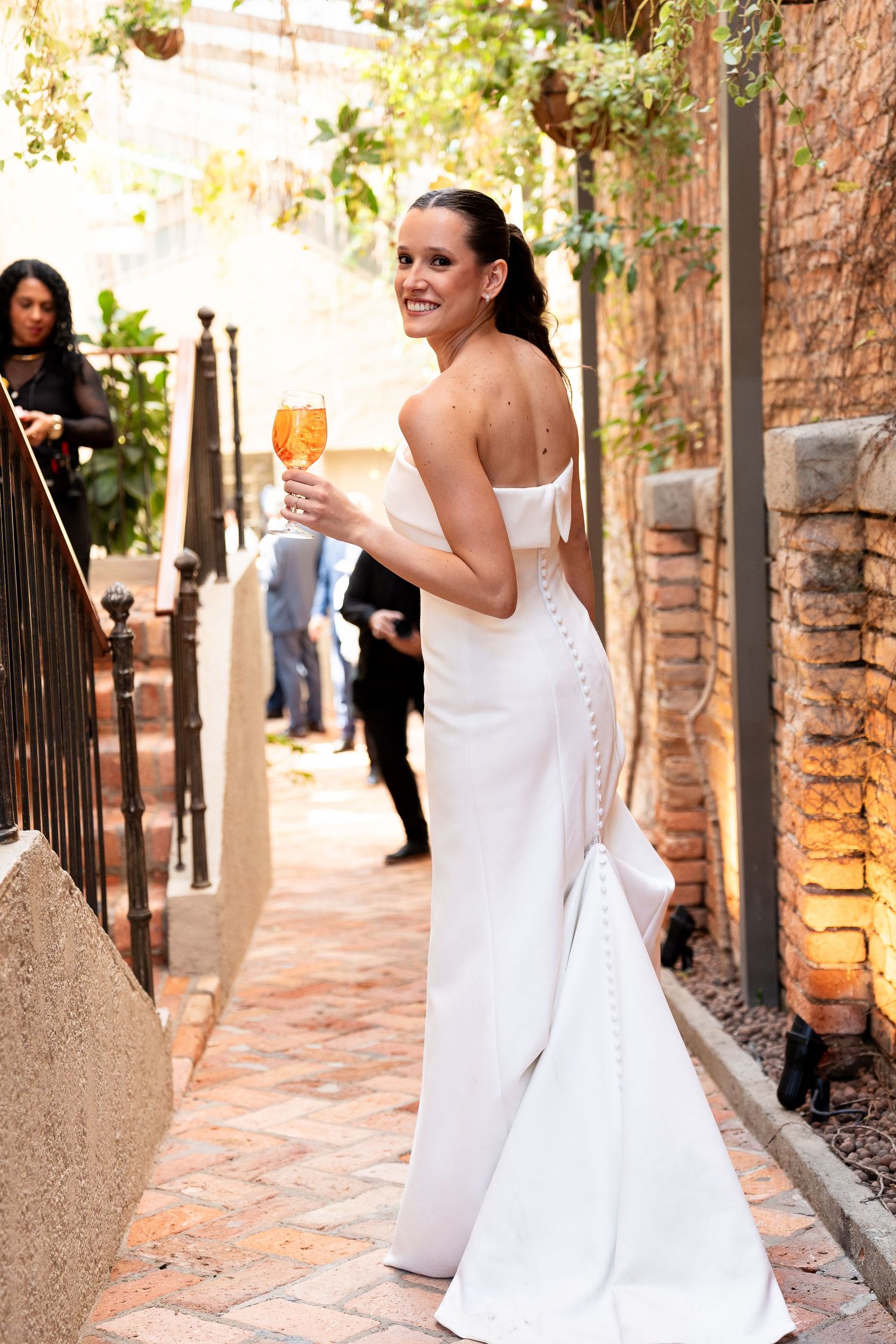 Bride in elegant white off-shoulder gown holding champagne glass, smiling outdoors on brick pathway.