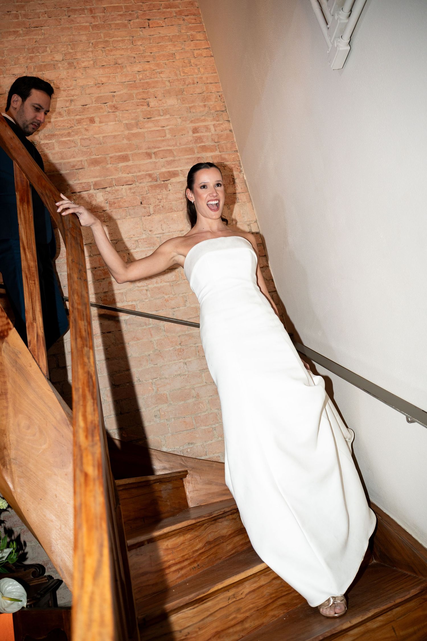Bride in white strapless gown laughing on wooden staircase with groom behind her near brick wall.