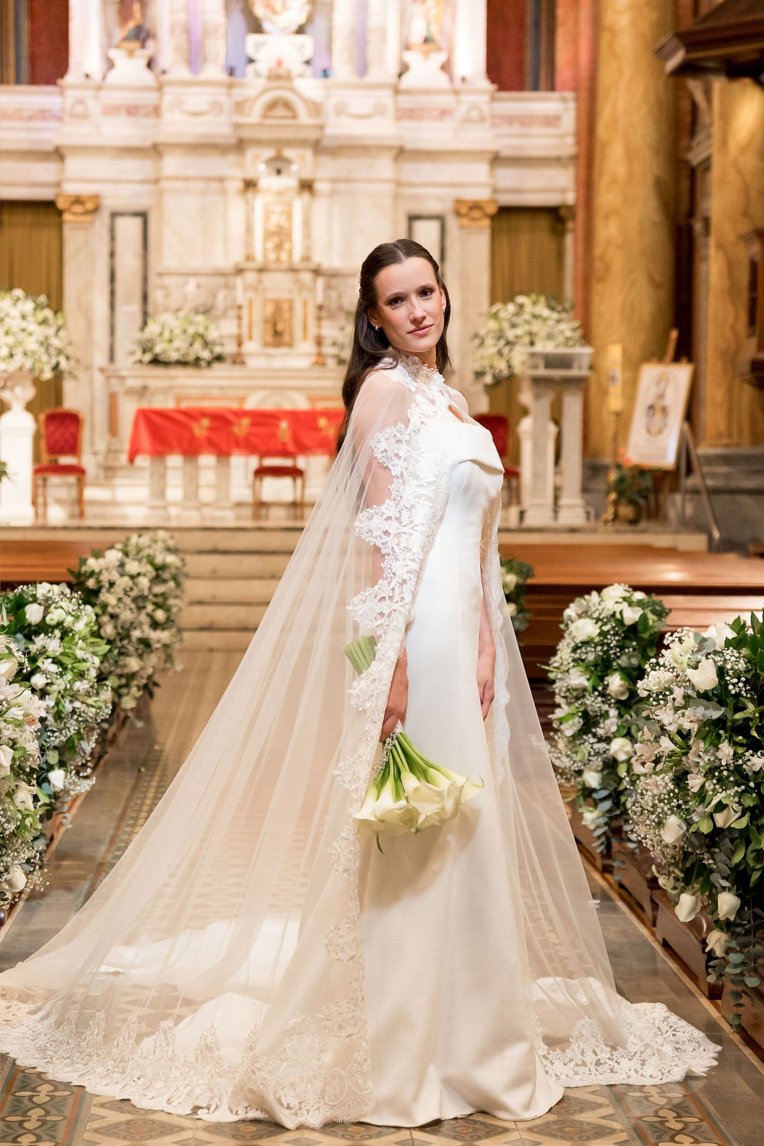 Bride in elegant white gown with lace cape veil holds calla lily bouquet in ornate church aisle with floral arrangements.