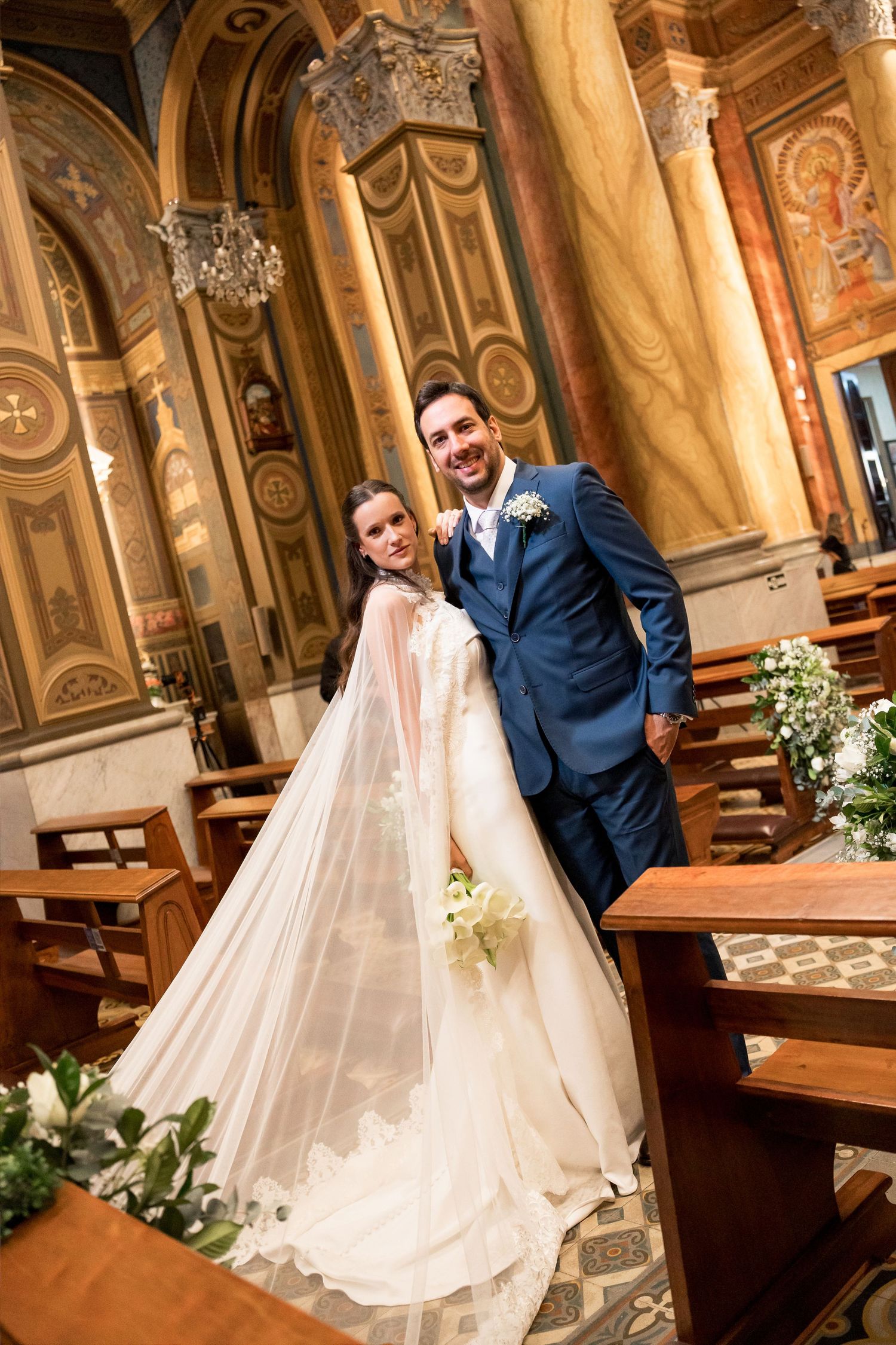 Bride and groom posing together inside an ornate church, bride wearing a flowing white gown with veil holding a bouquet.