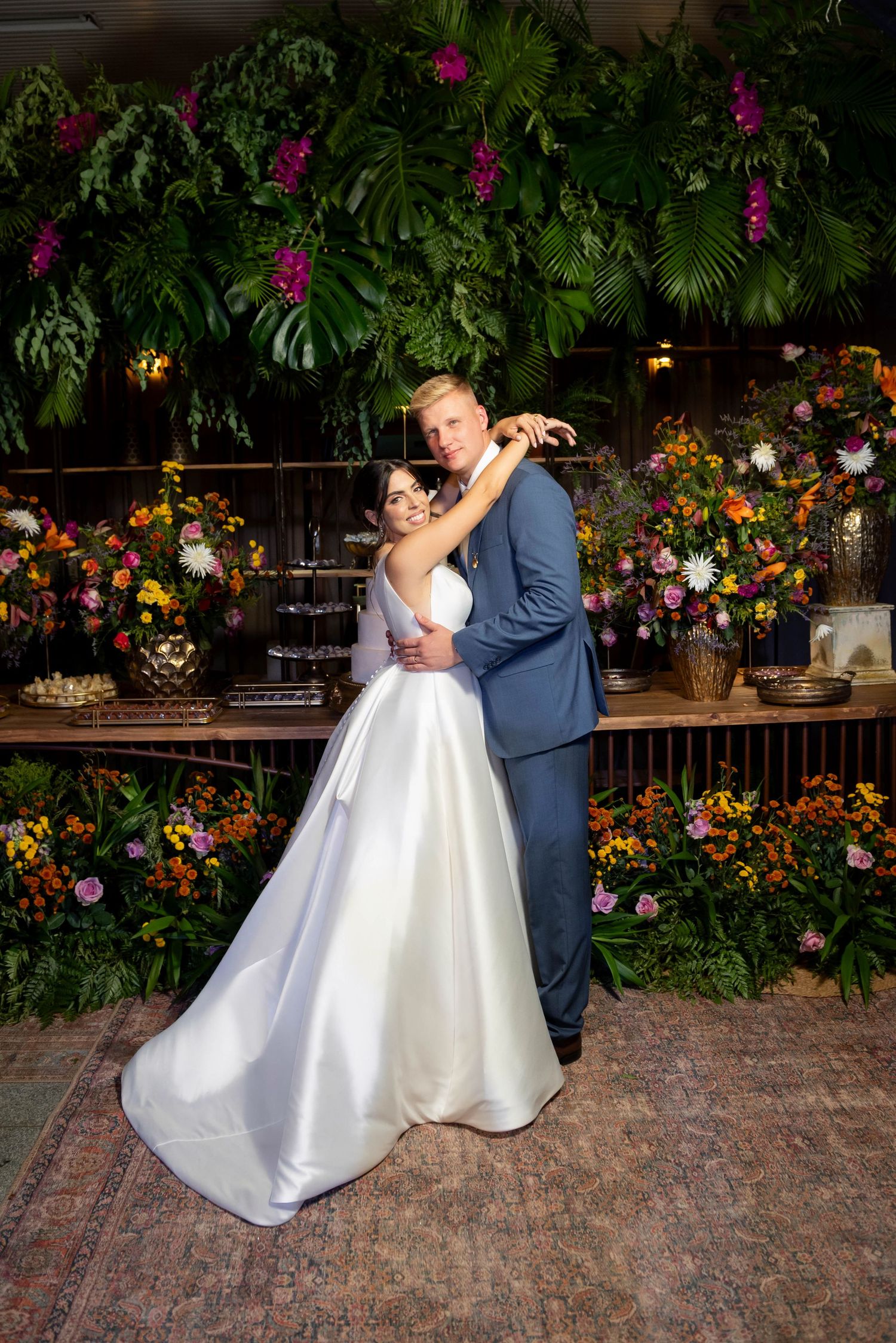 Bride in white gown and groom in blue suit embrace in front of lush tropical greenery and colorful floral arrangements.