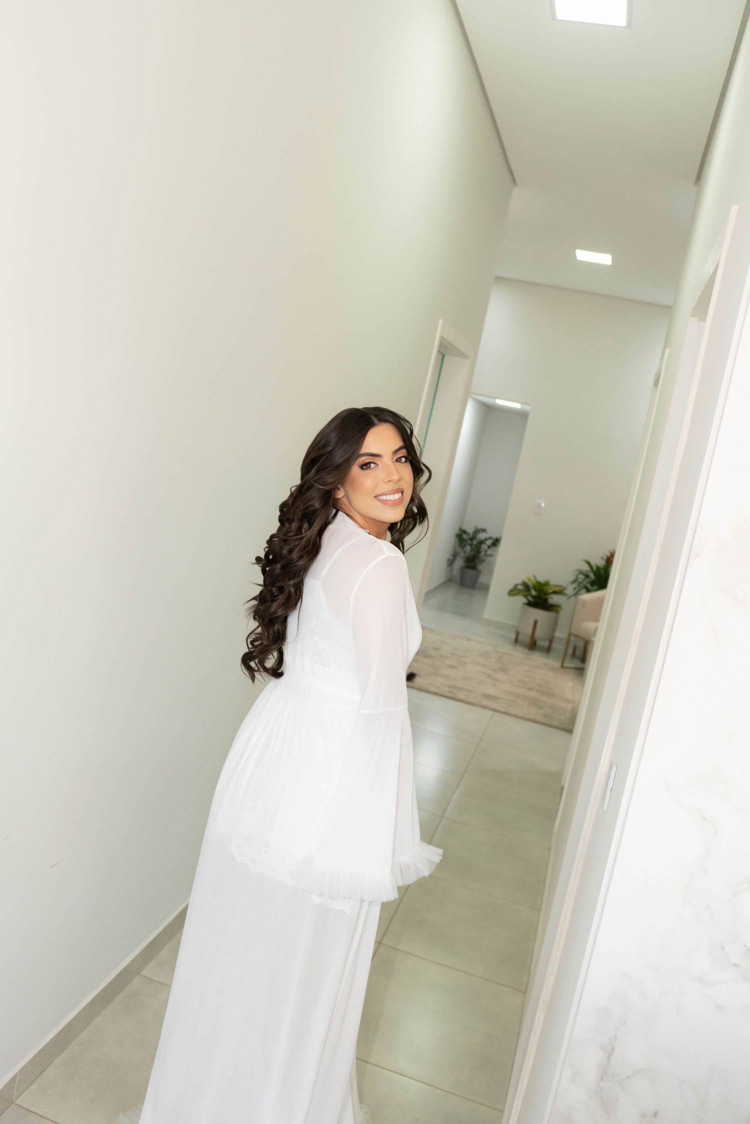 A smiling brunette bride in a flowing white dress and robe poses gracefully in a bright modern hallway.