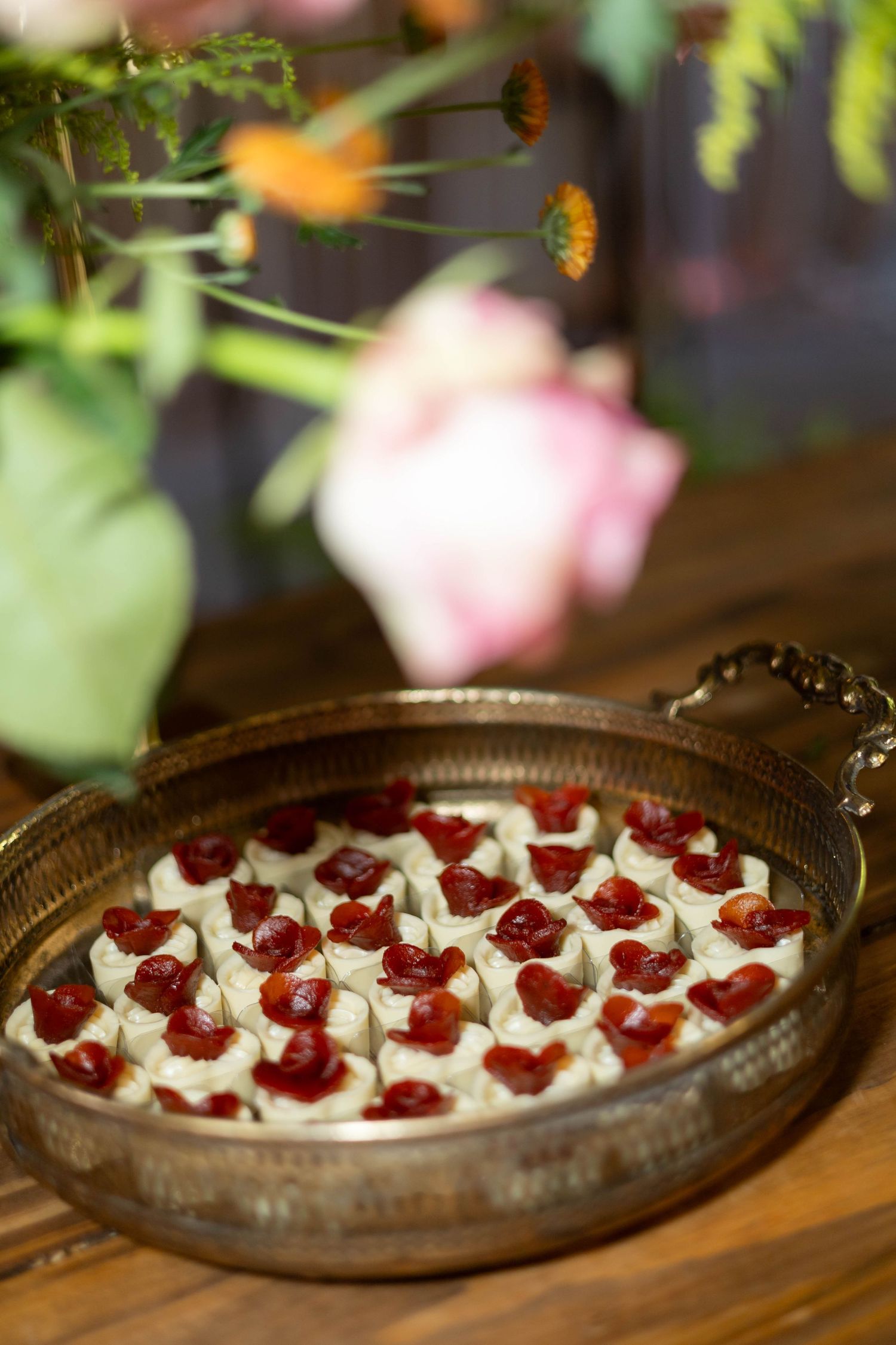Elegant silver tray of cherry topped cream appetizers displayed on wooden table with floral arrangement in background.