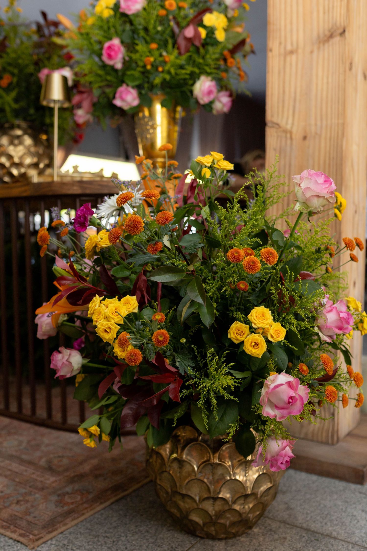 Lush floral arrangement in gold vase with pink roses, yellow blooms, and orange flowers displayed indoors.