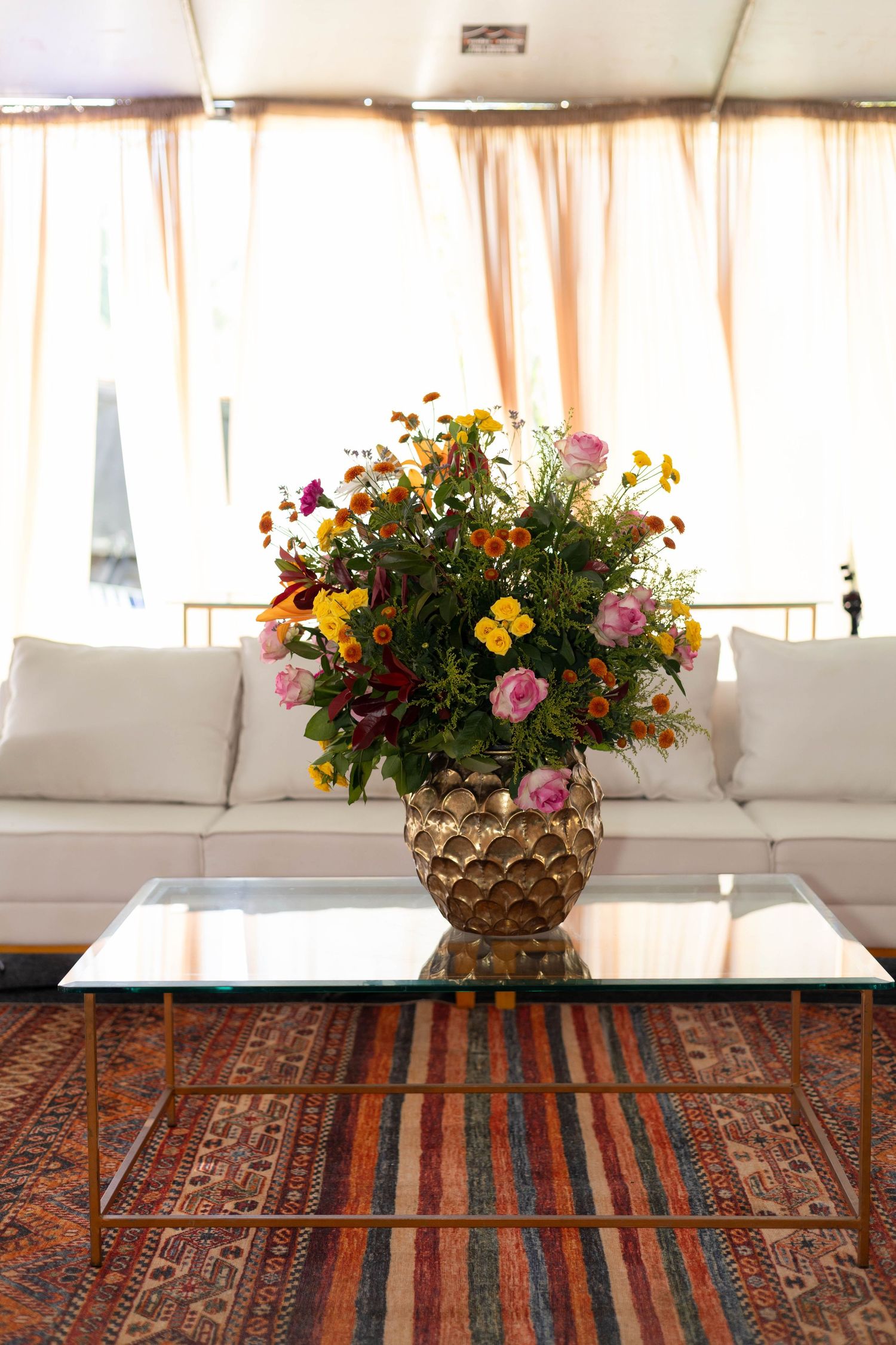Gold vase with colorful wildflowers on glass coffee table in elegant white living room with patterned rug.