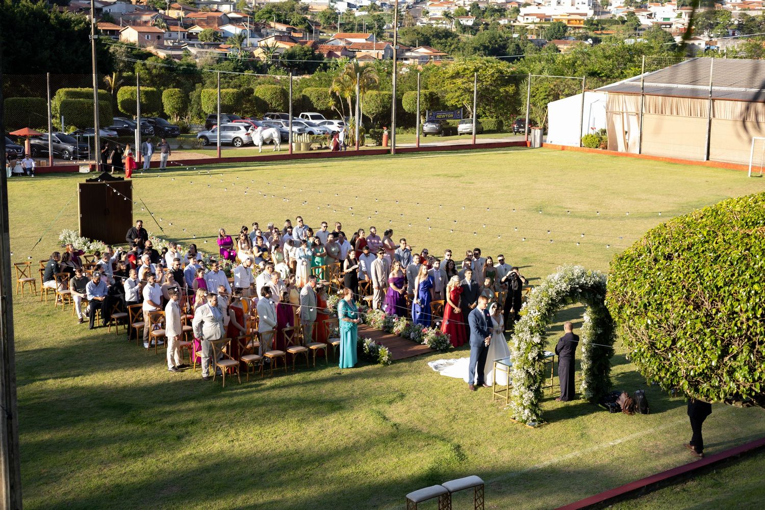 Outdoor wedding ceremony on a lush green lawn with guests seated in rows facing the couple at a floral arch.
