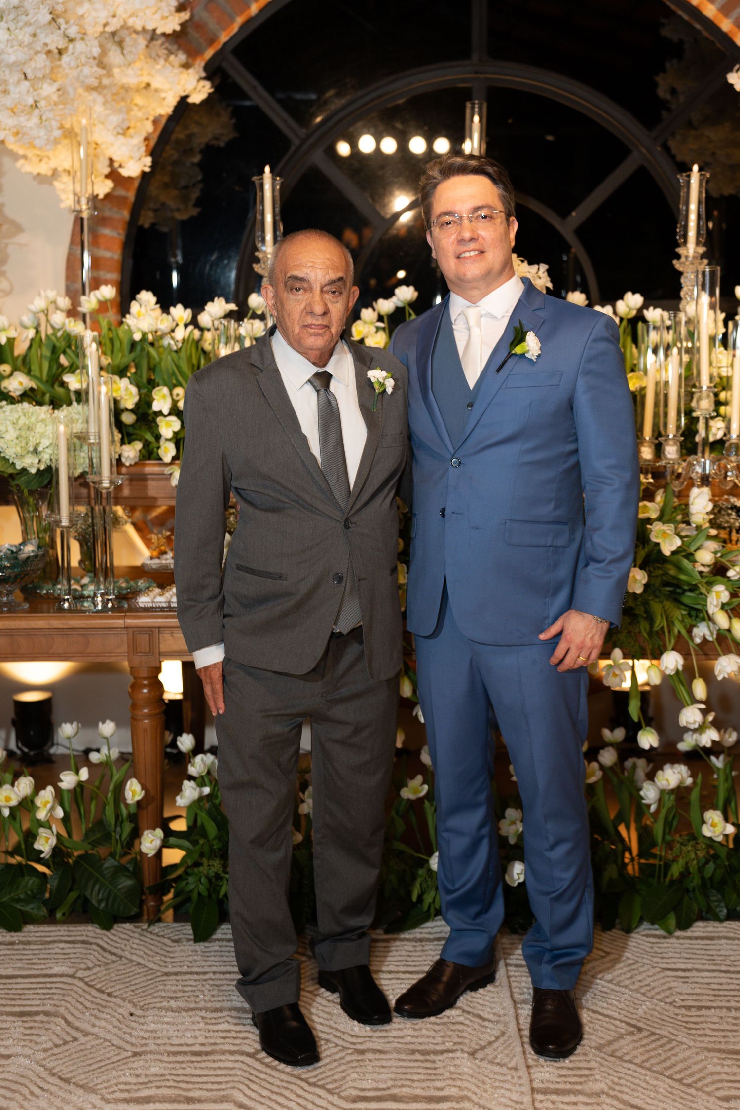Two men in suits pose together at an elegant wedding reception with white floral arrangements.