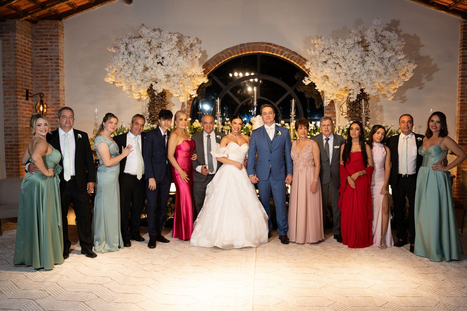 Wedding party poses together in elegant venue with floral arch, bride in white gown surrounded by guests in formal attire.