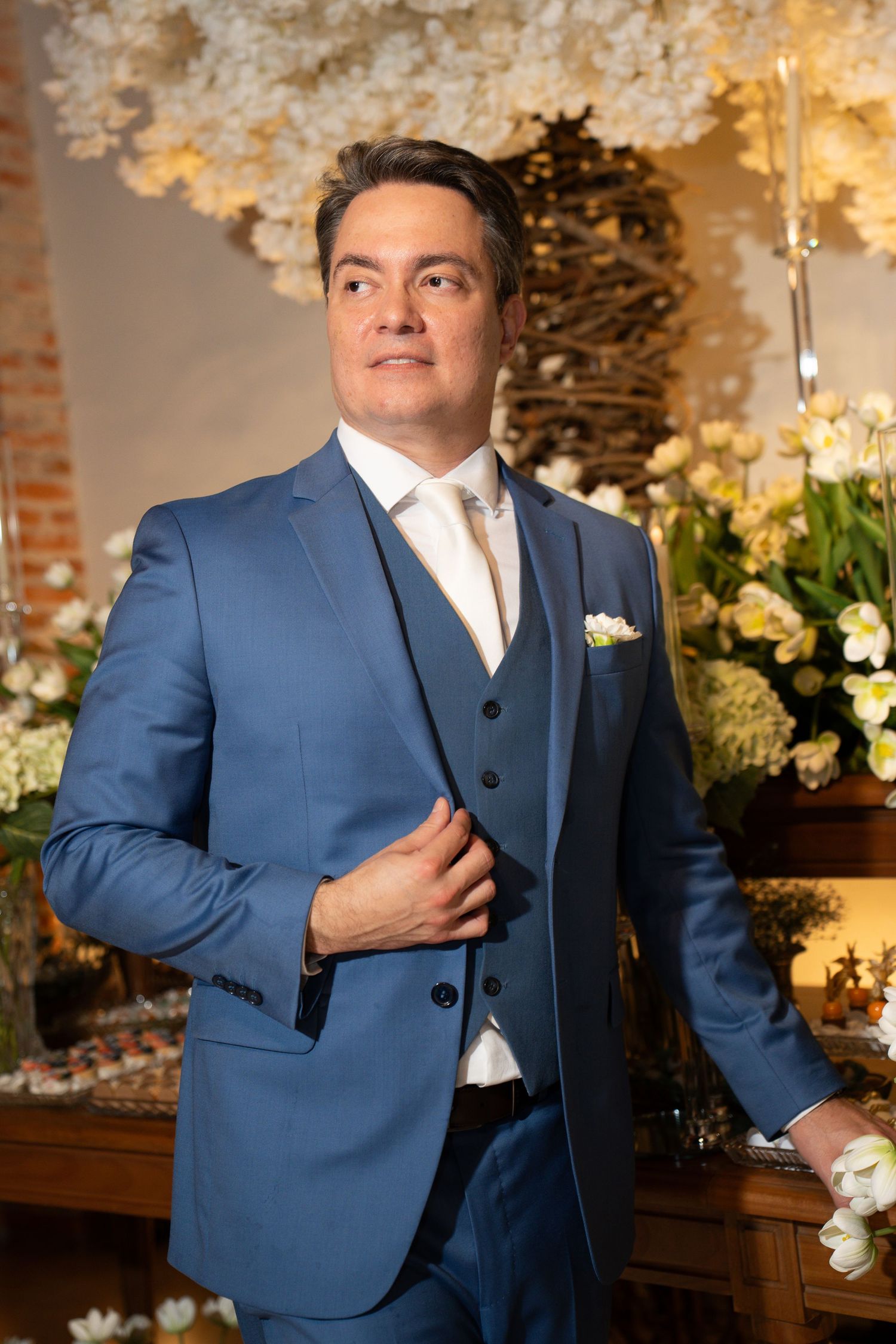 A man in a blue three-piece suit with white boutonniere poses elegantly before a floral backdrop at a formal event.