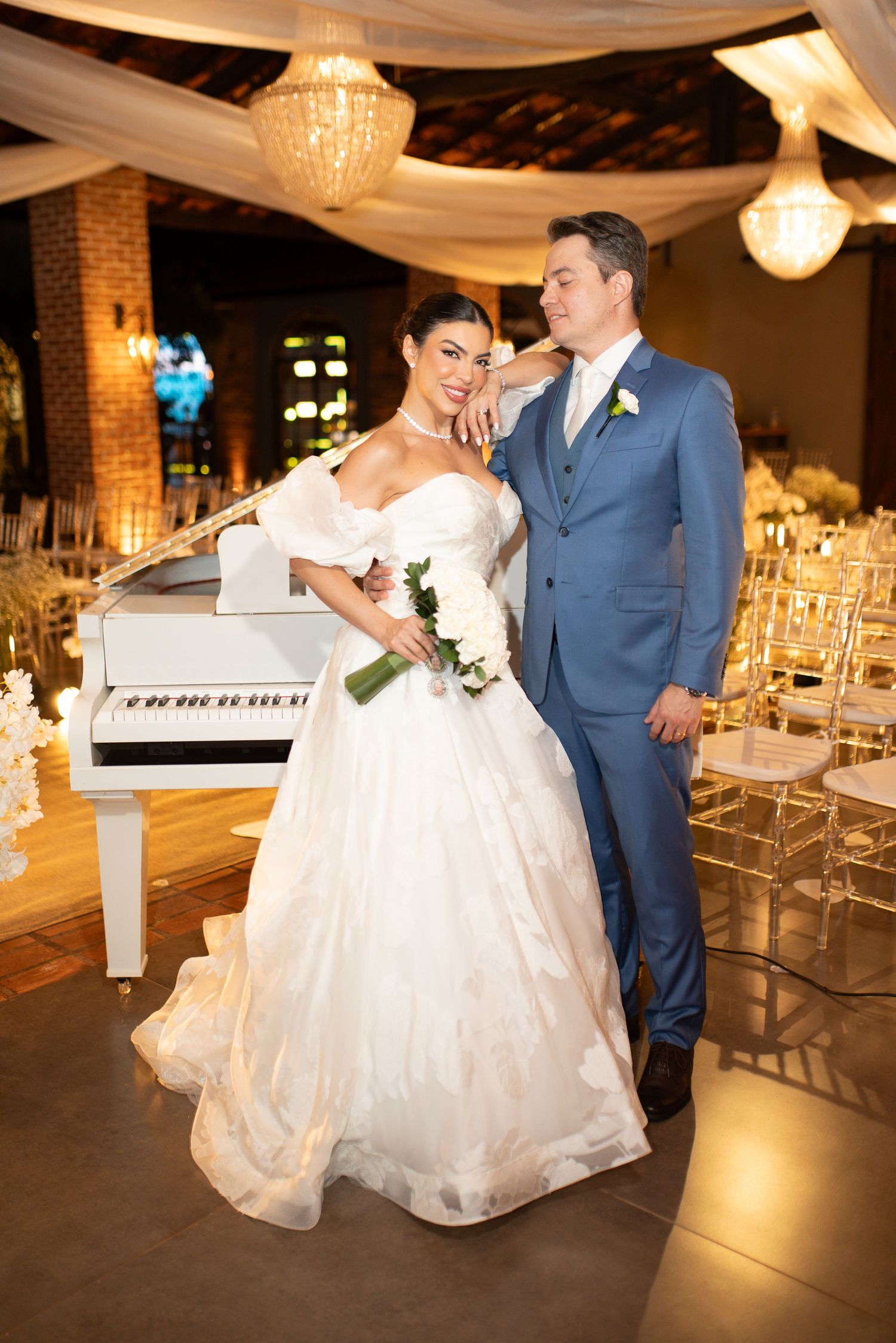 Bride in white ballgown and groom in blue suit pose together near a white grand piano at elegant wedding venue.