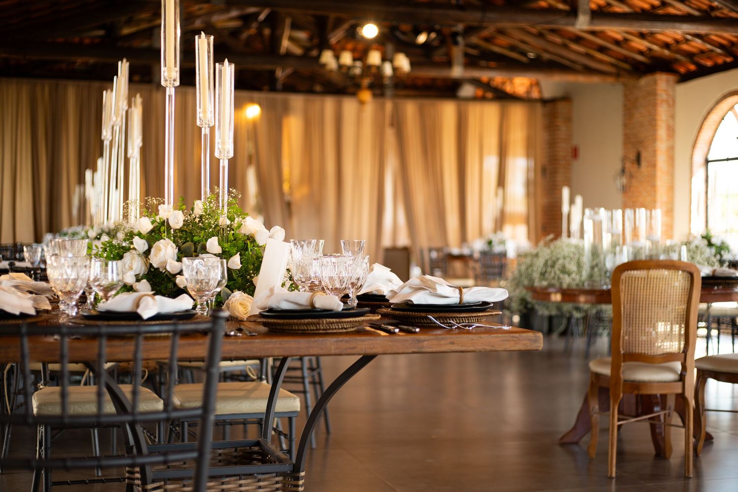 Elegant wedding reception hall with wooden tables, white floral centerpieces, tall candles, and gold chairs.