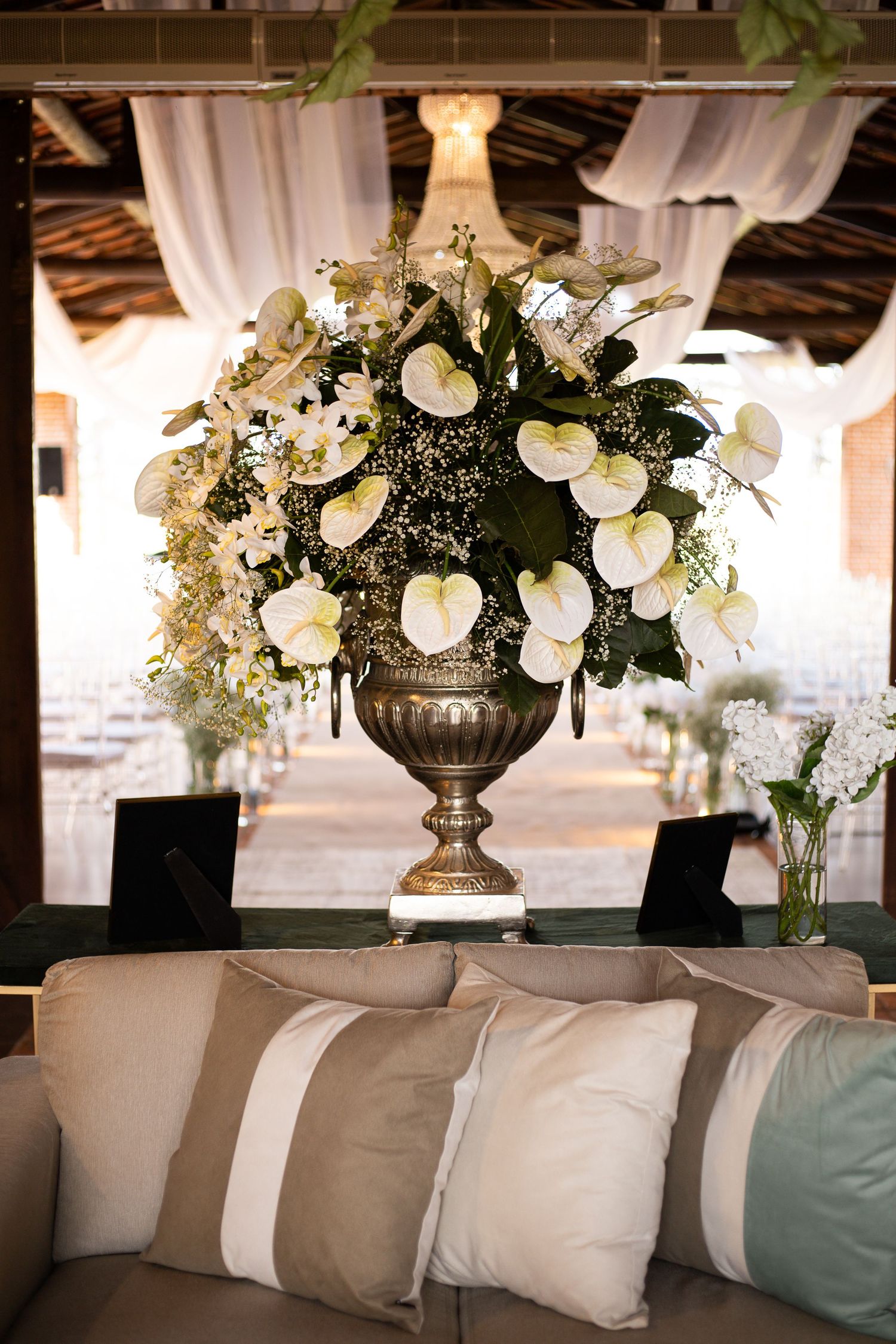 Elegant urn vase with white anthurium and orchid floral arrangement on a sofa table in a decorated event venue.