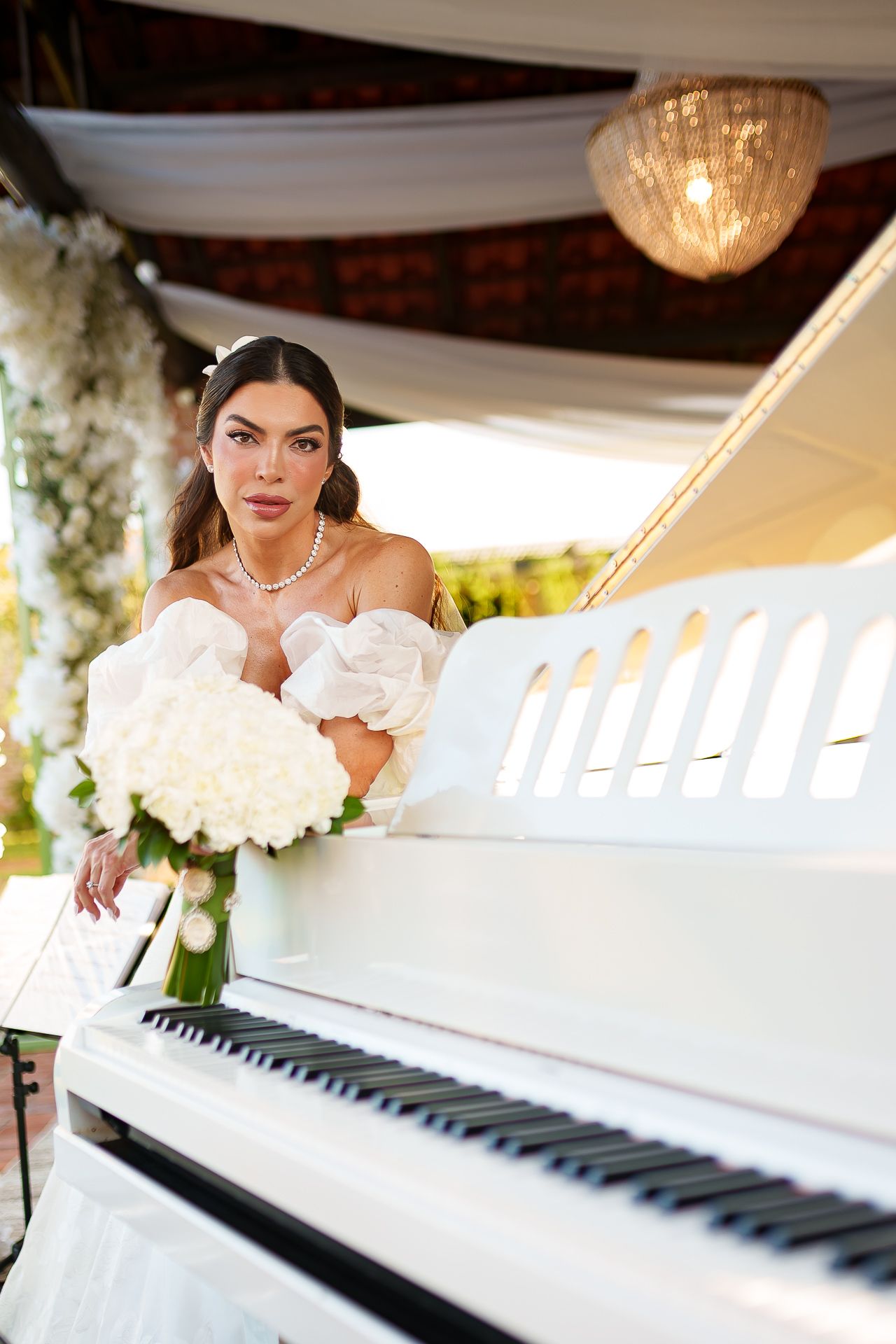 Elegant bride in white off-shoulder gown holding white rose bouquet, leaning on a white grand piano at outdoor wedding venue.