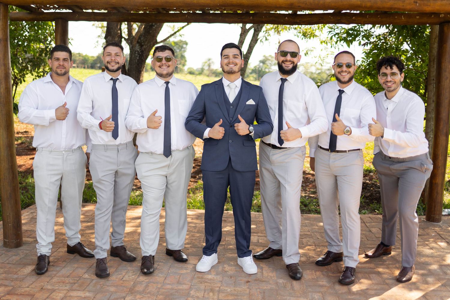 Groom in navy suit poses with six groomsmen in white shirts and ties, giving thumbs up outdoors at wedding venue.