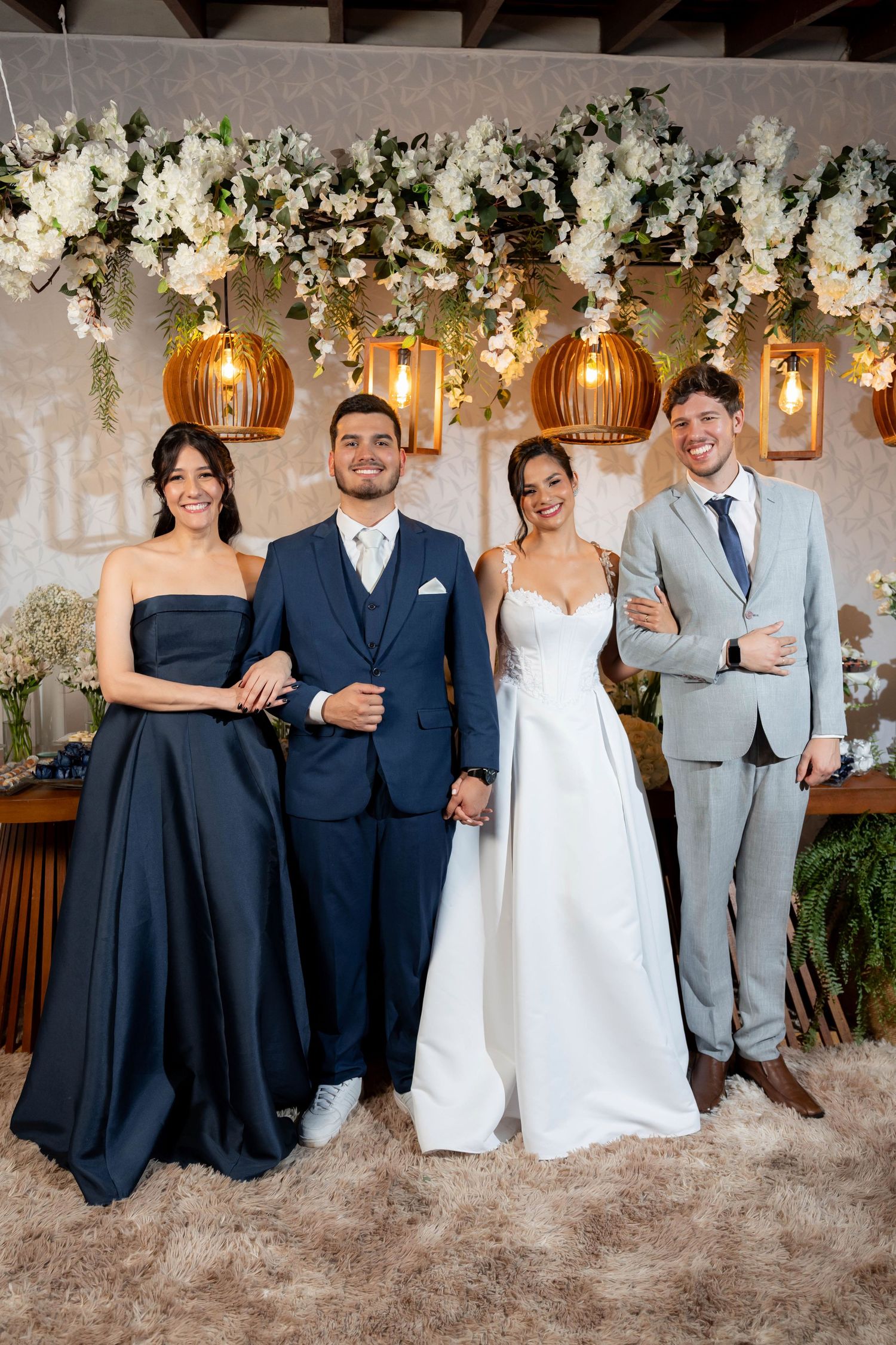 Wedding couple smiles with two guests under white floral arch with warm golden hanging lanterns.