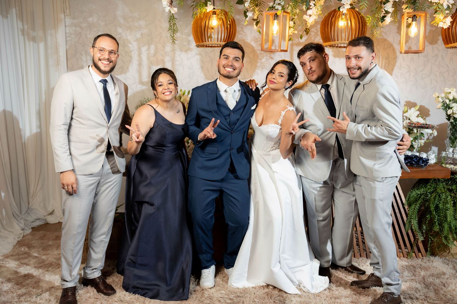 Wedding party poses joyfully in elegant venue, bride in white gown with groom in navy suit surrounded by smiling guests.