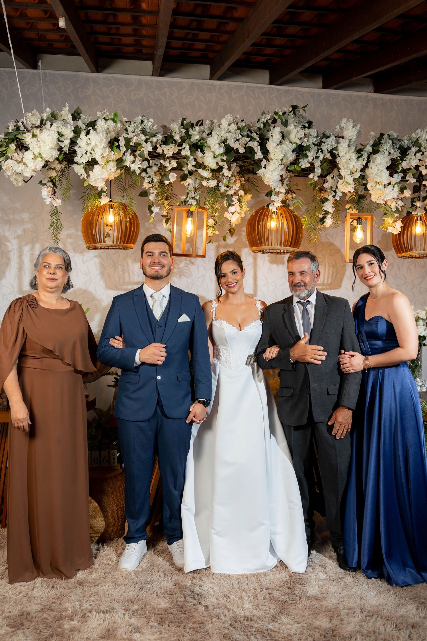Wedding party of five posing under floral arch with pendant lights, bride in white gown flanked by groom and family.