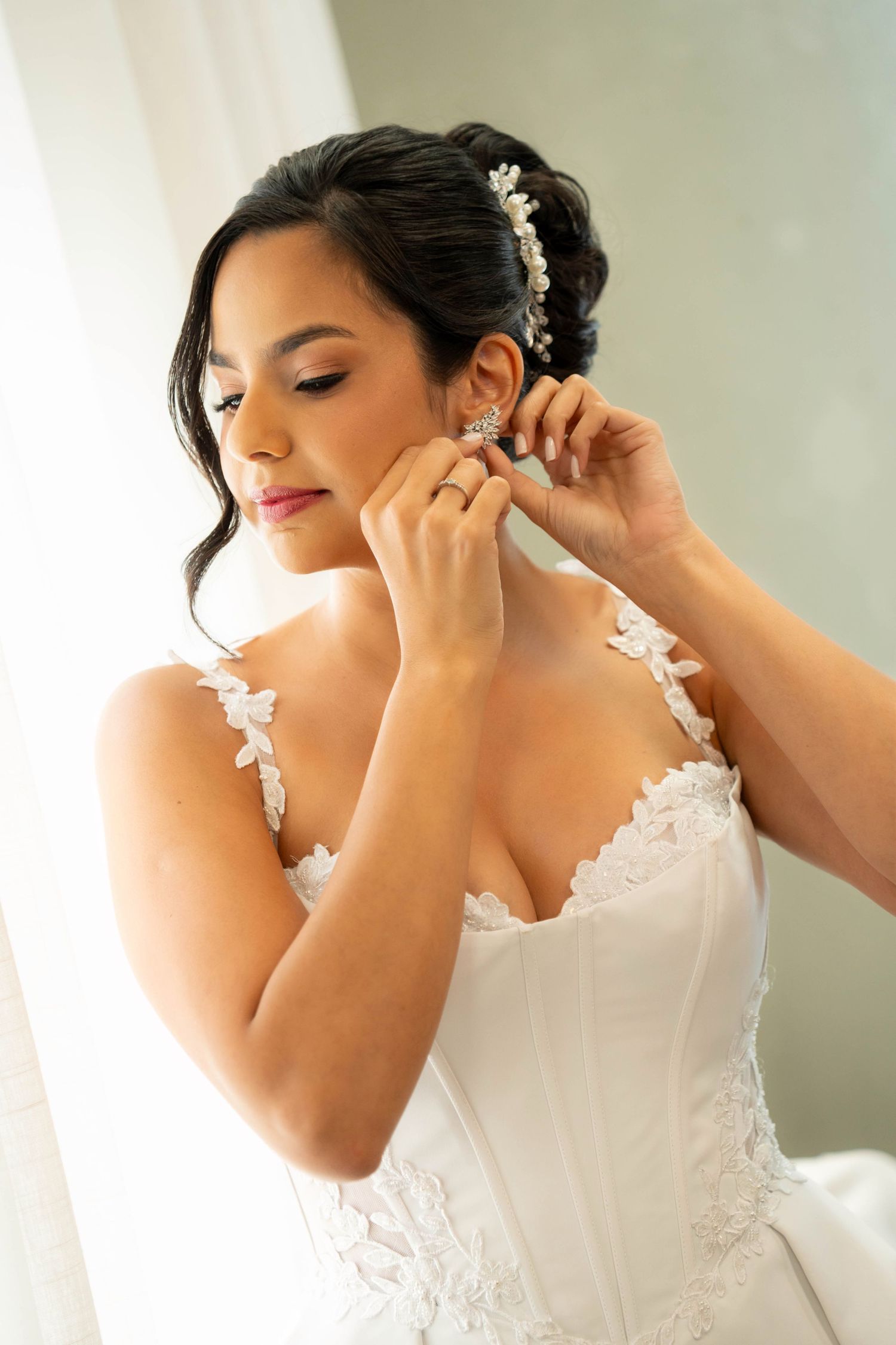 Bride in white lace wedding gown putting on pearl earrings with floral hair comb accessory.