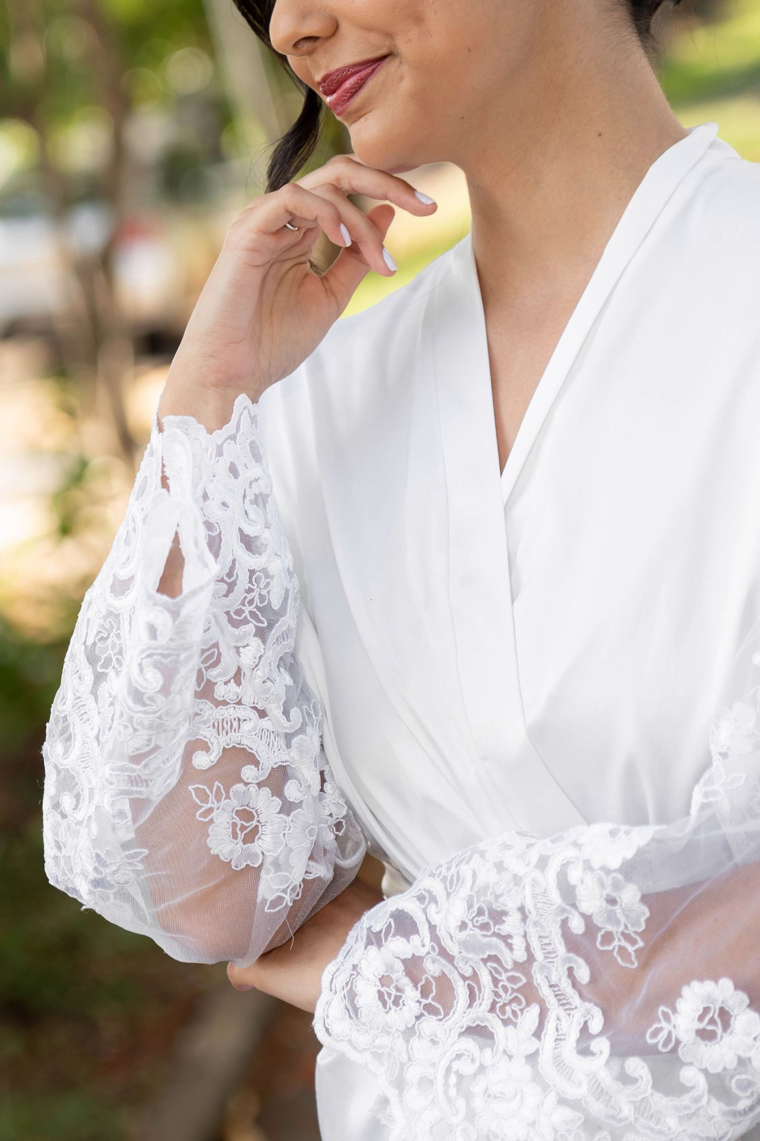 Woman in white lace bridal robe with elegant floral embroidery on sleeves, smiling softly outdoors.