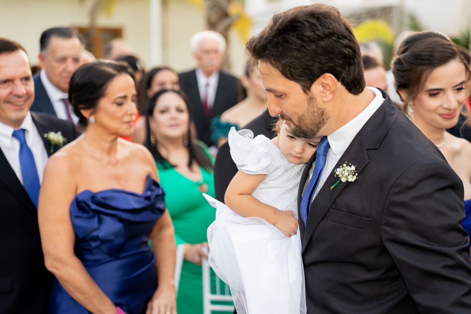 Groom in navy suit tenderly holds a young girl in white dress during an outdoor wedding ceremony with guests watching.