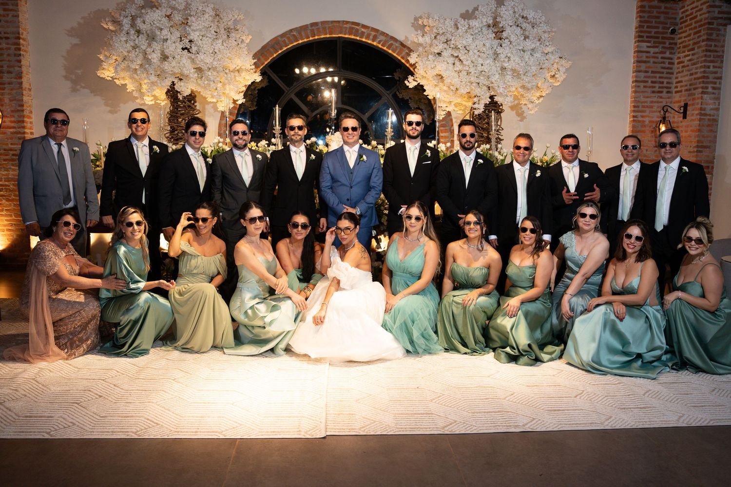 Wedding party in elegant venue, bridesmaids in sage green gowns and groomsmen in suits, all wearing sunglasses.