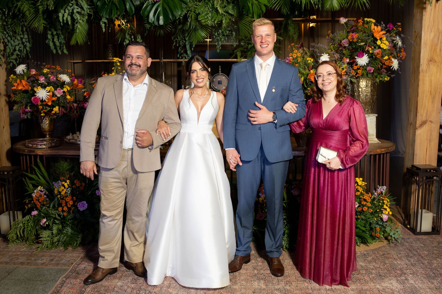 Wedding party of four posing in front of lush floral backdrop, bride in white gown with groom in blue suit.