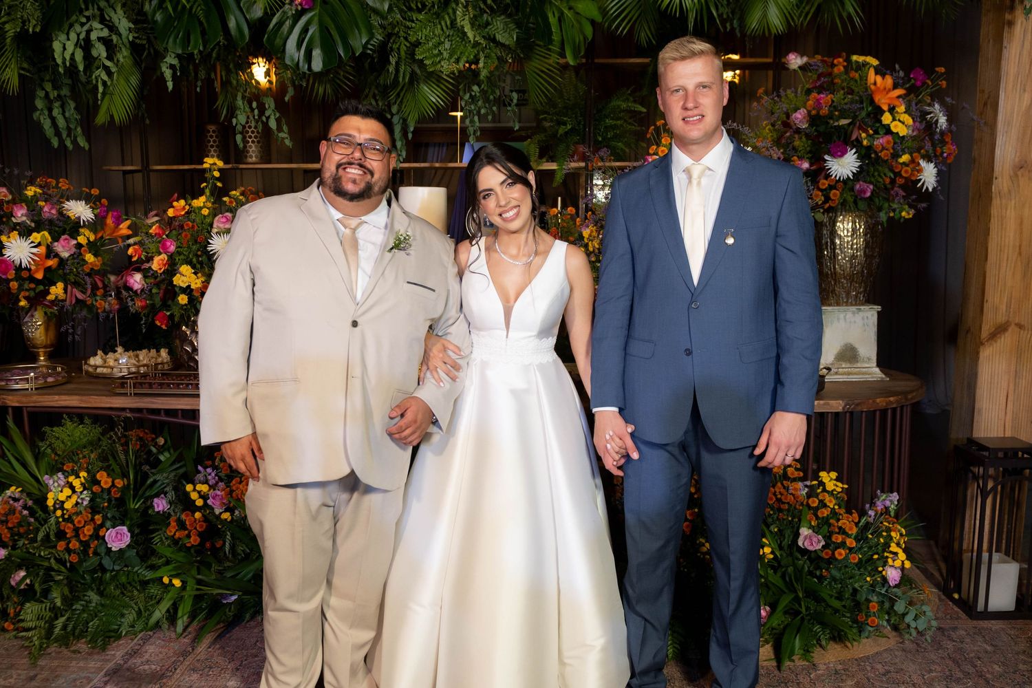 Bride in white gown poses with two men in suits at a floral-decorated wedding reception venue.