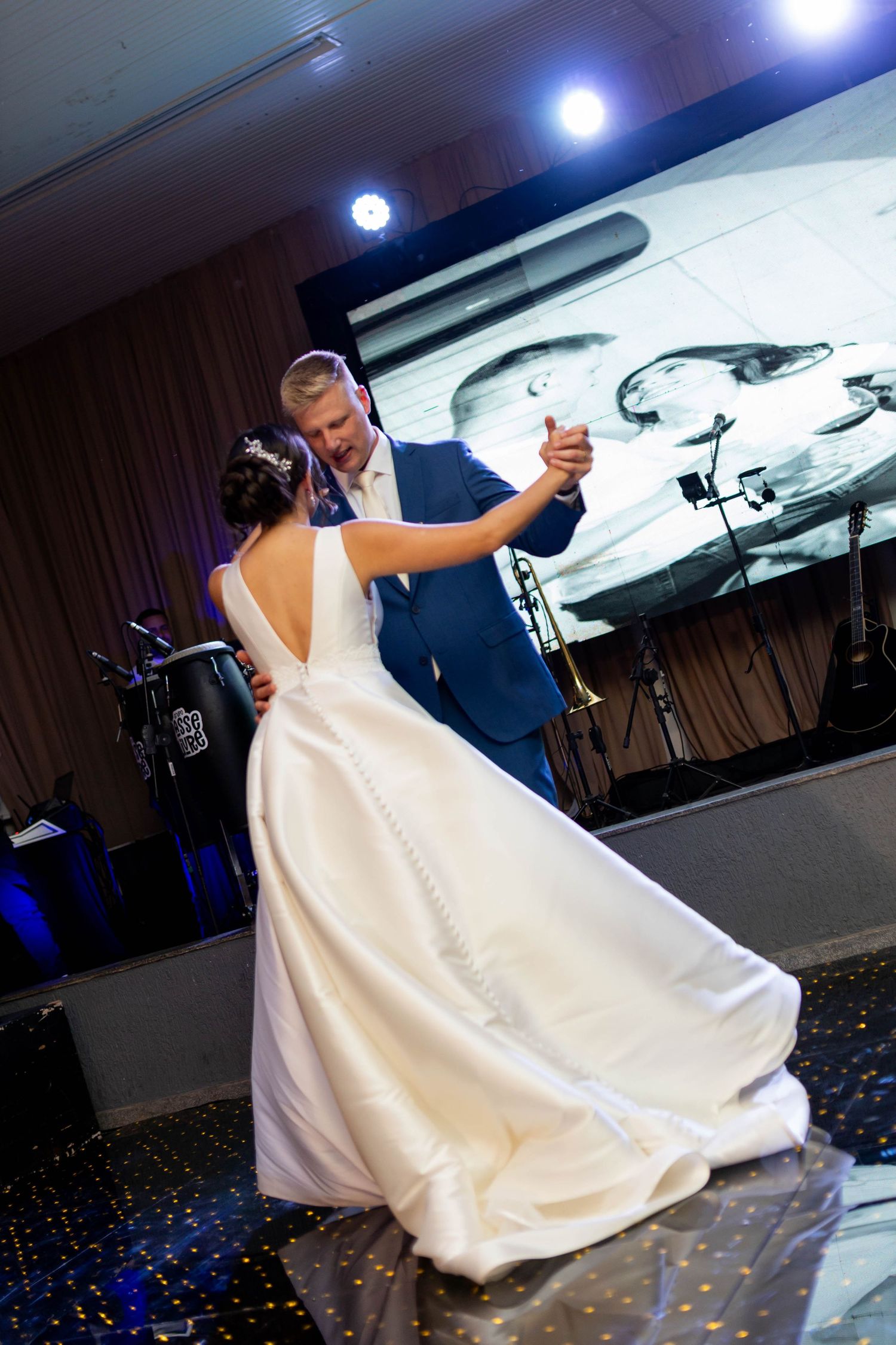 Bride in white gown and groom in blue suit share their first dance on a sparkling dance floor at wedding reception.