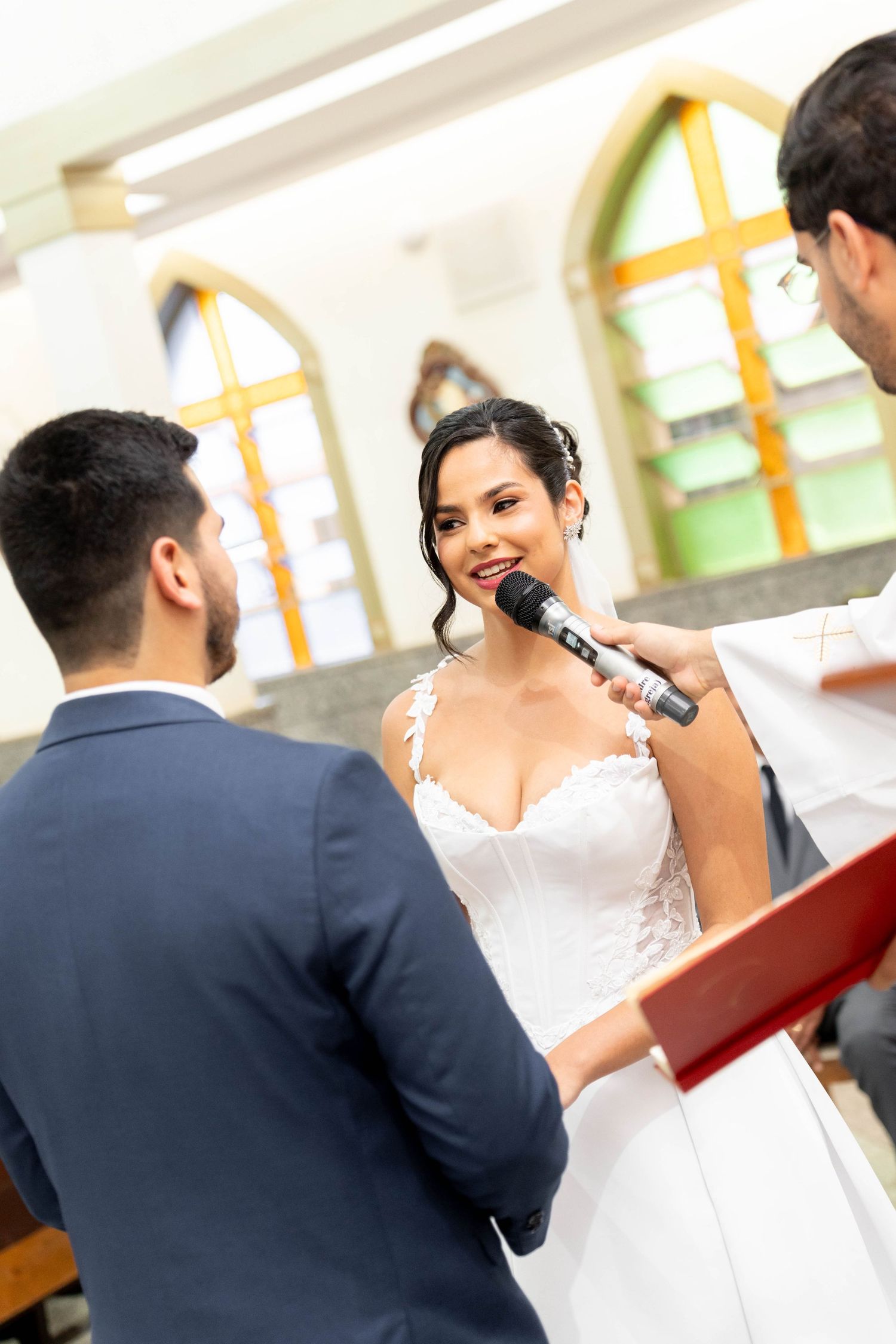 Bride smiling while reciting wedding vows at church altar, groom facing her as officiant holds microphone.
