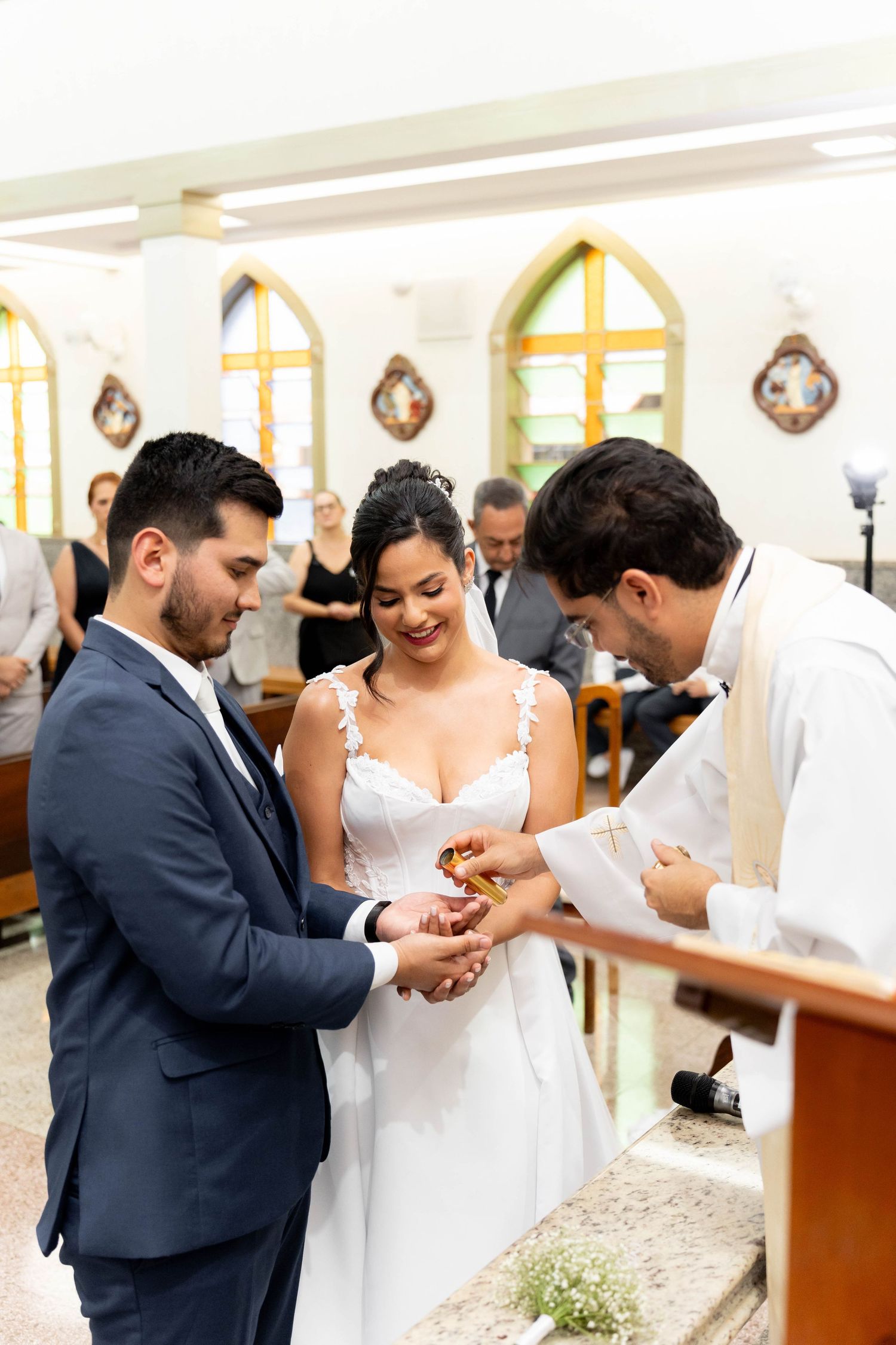 Bride and groom exchange rings during a church wedding ceremony, officiated by a priest in white robes.