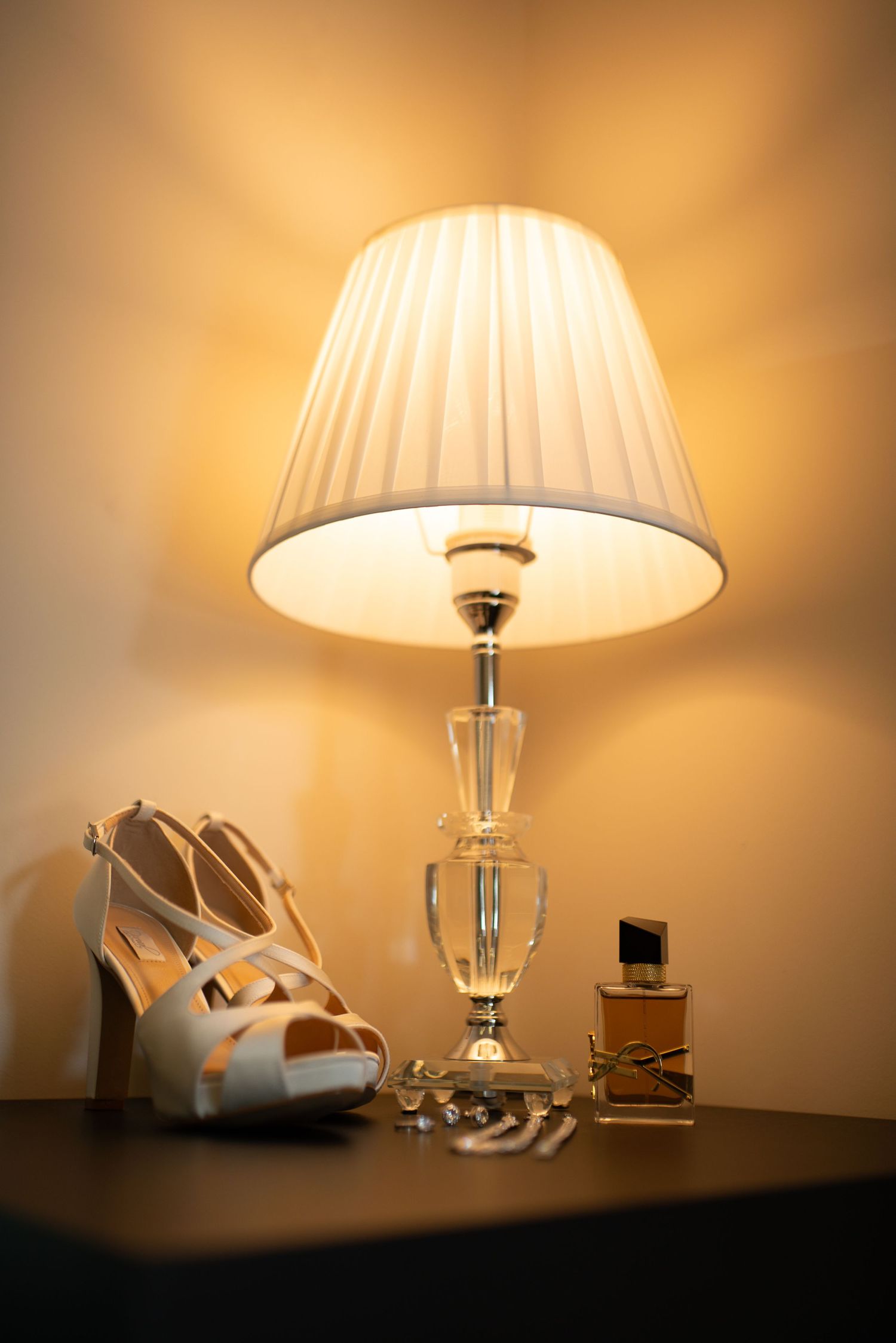 Elegant table lamp with crystal base illuminating white strappy heels and YSL perfume bottle on a dark nightstand.