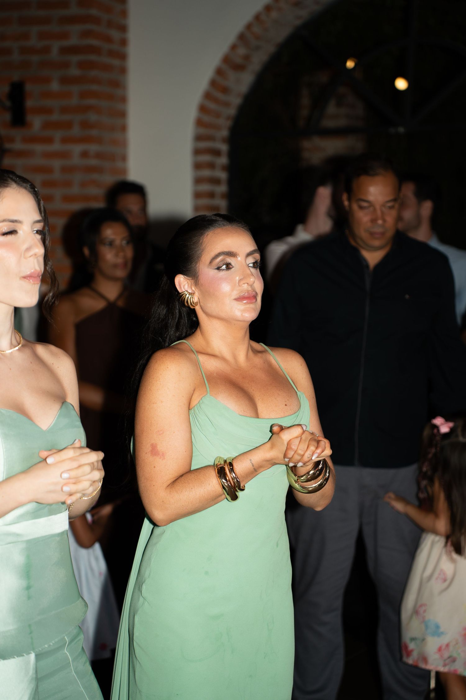 Two women in sage green satin dresses dancing at an elegant indoor event with brick arch backdrop.