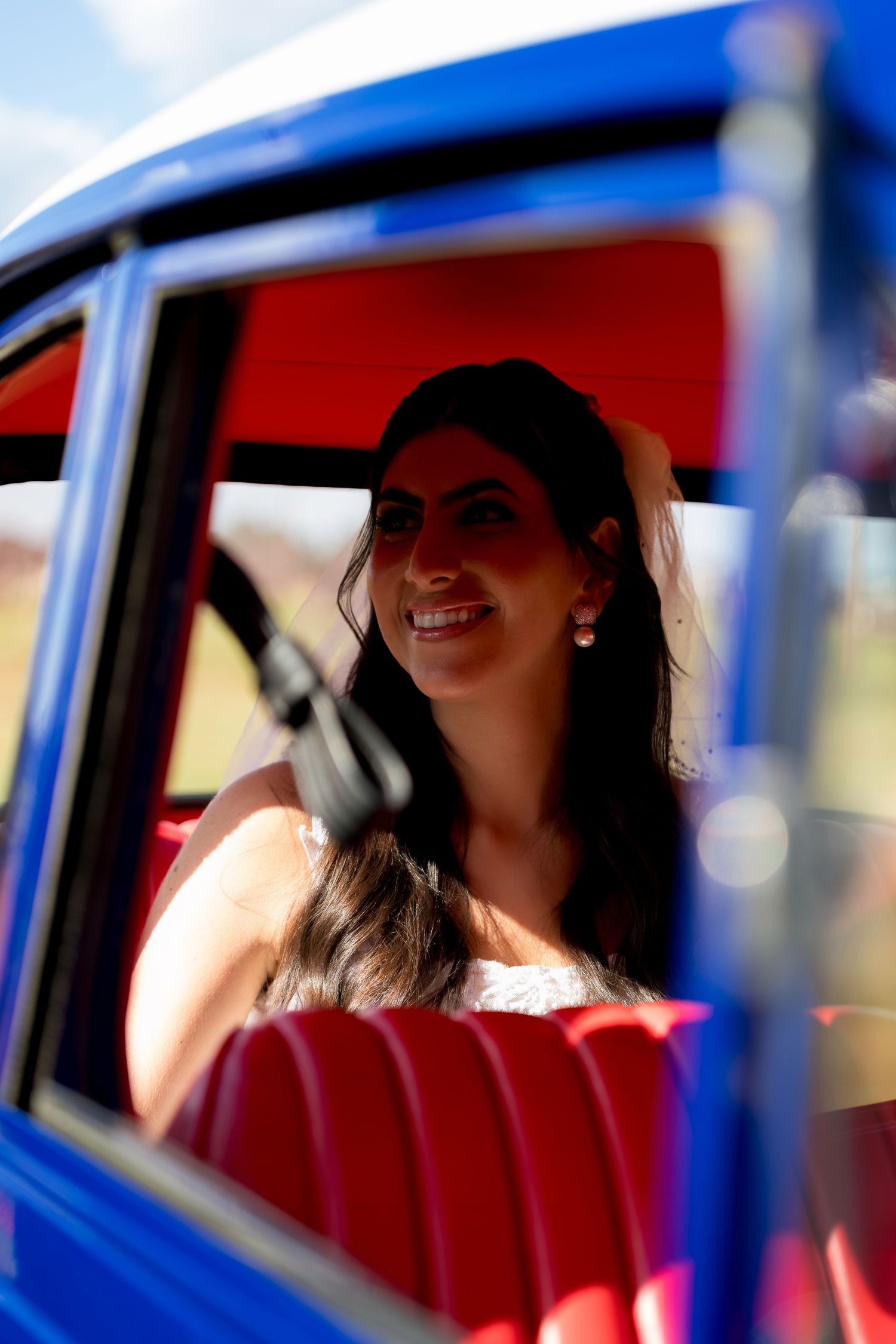 Smiling bride with dark hair sitting in a vintage blue car with red interior on a sunny day.