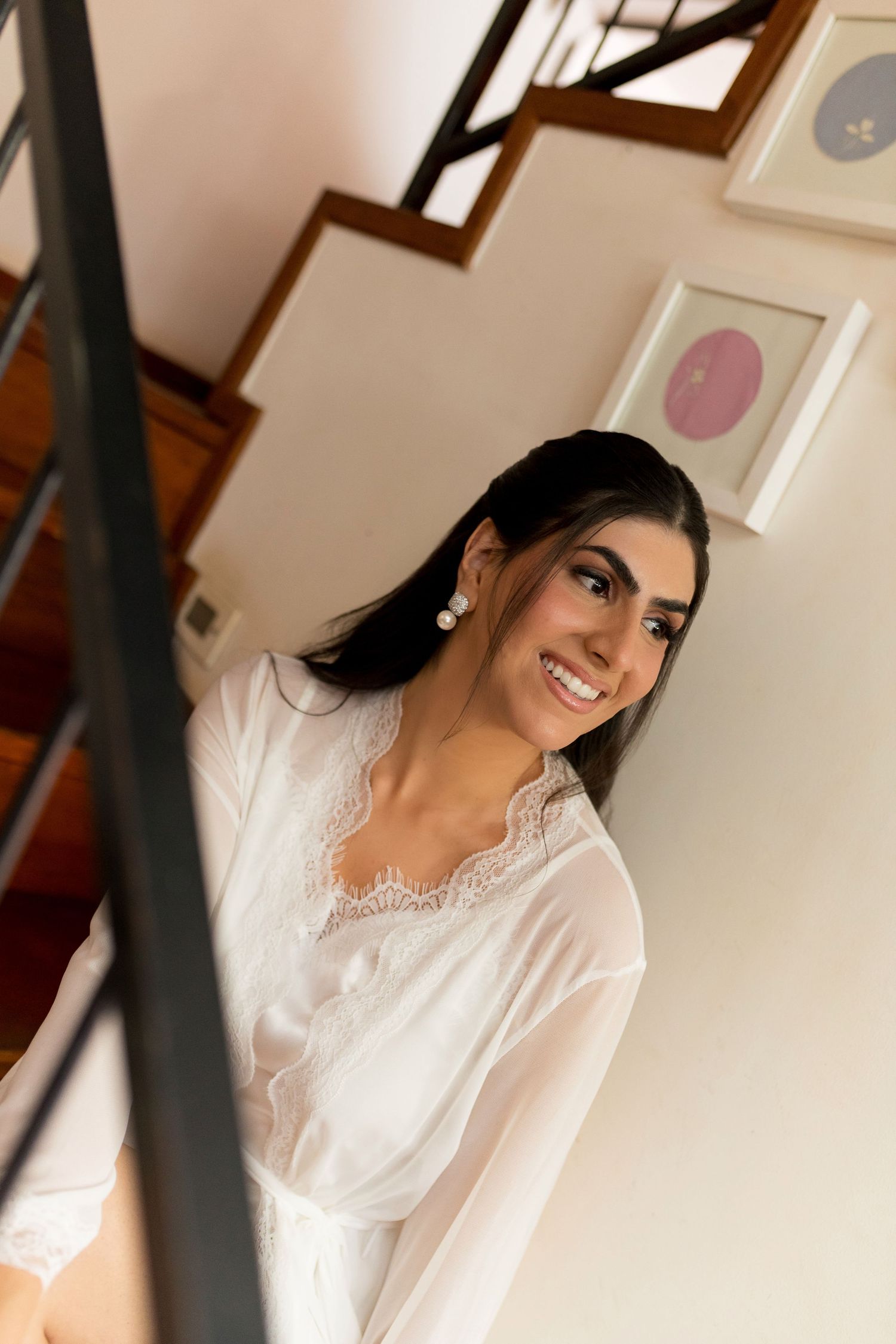 Smiling young woman in white lace robe standing on staircase with framed art on wall behind her.