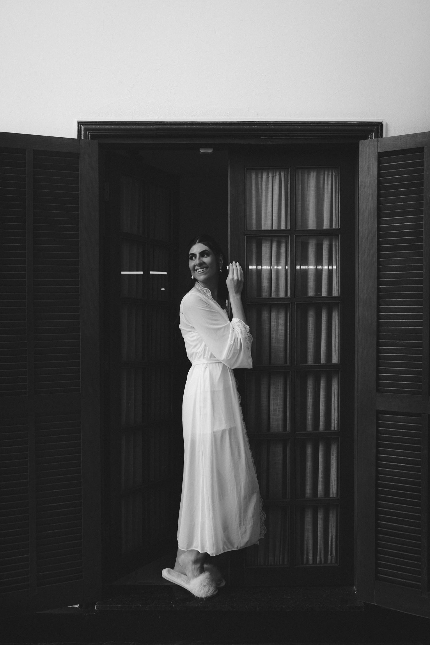 Black and white photo of a smiling woman in a white robe standing in an open doorway with shutters.