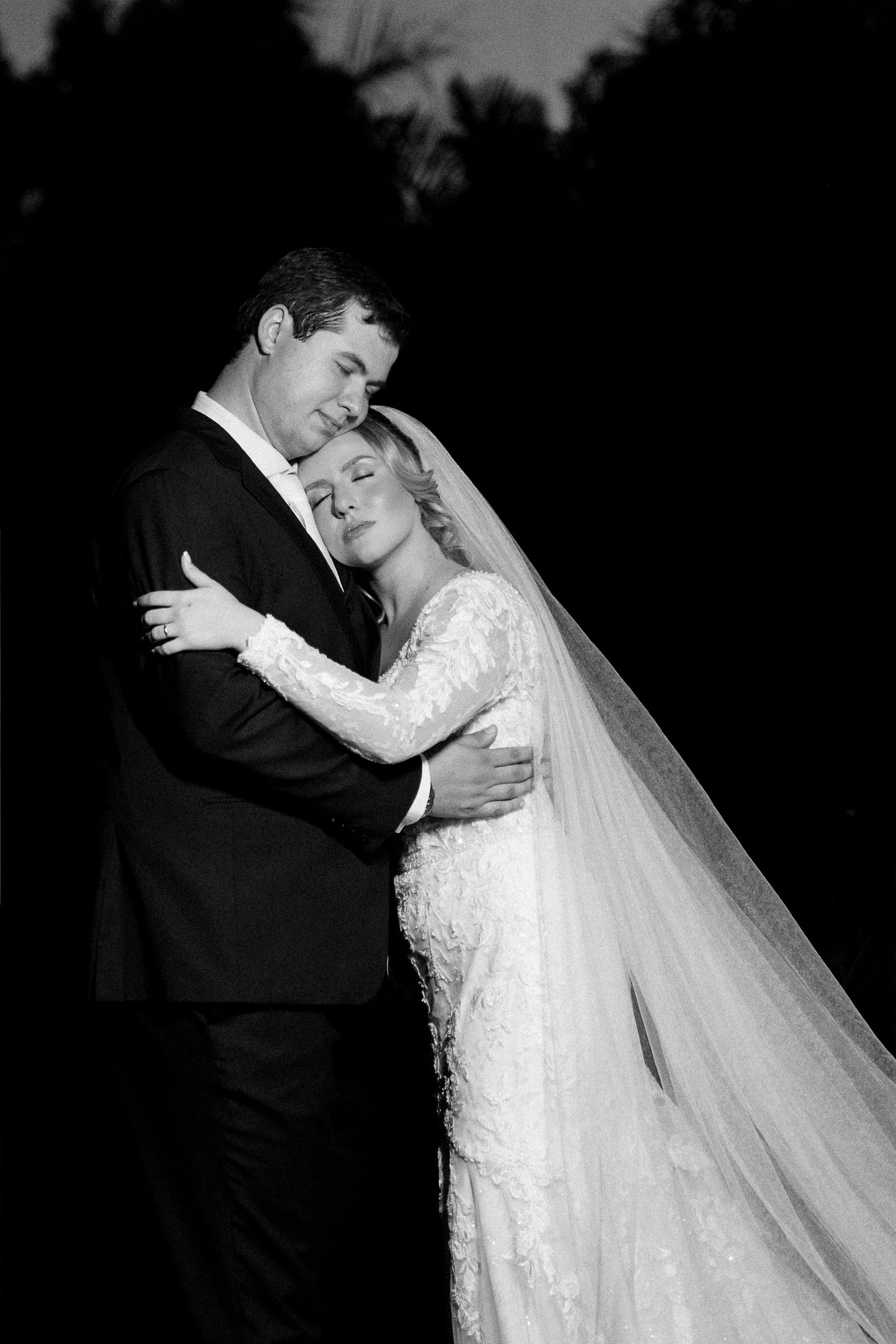 Black and white wedding portrait of bride and groom embracing tenderly at night, bride in lace gown and veil.
