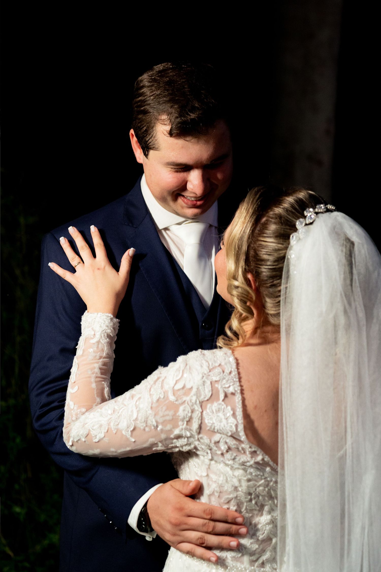 Bride in lace gown shows ring while groom in navy suit smiles, sharing a romantic wedding moment.