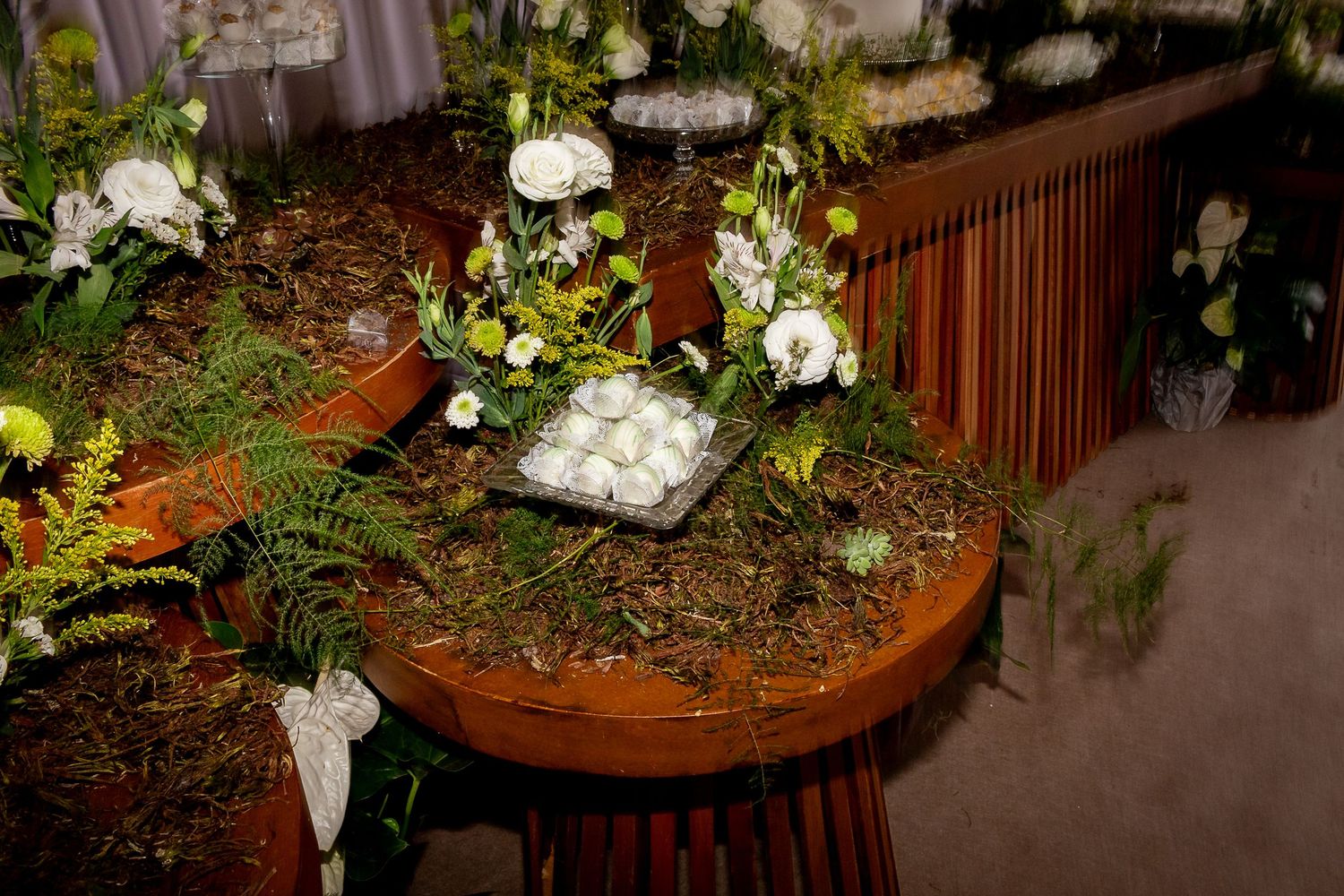 Rustic wooden table decorated with lush ferns, white flowers, and a tray of white treats for an elegant event.