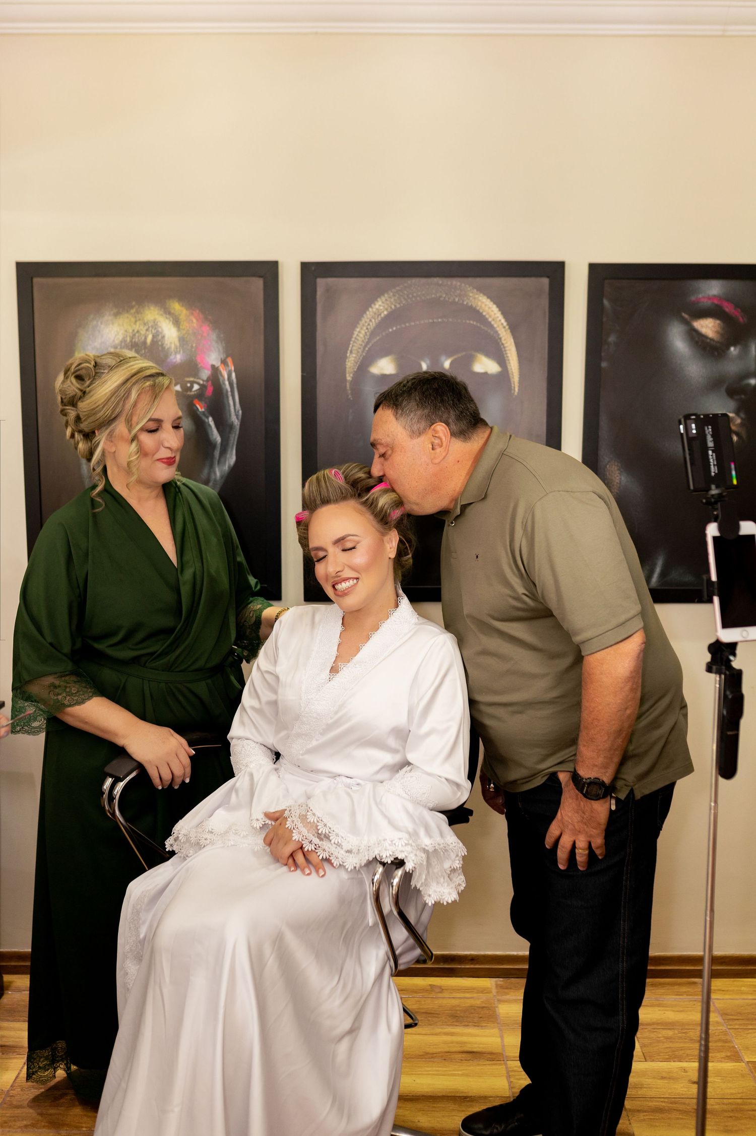 A bride in white robe sits smiling as a man kisses her cheek while a woman in green dress looks on.