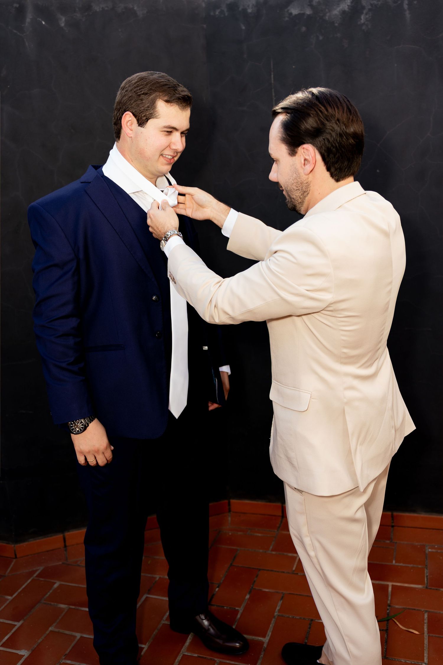 A man in a cream suit adjusts the white tie of a groom in a navy blue suit before a wedding ceremony.