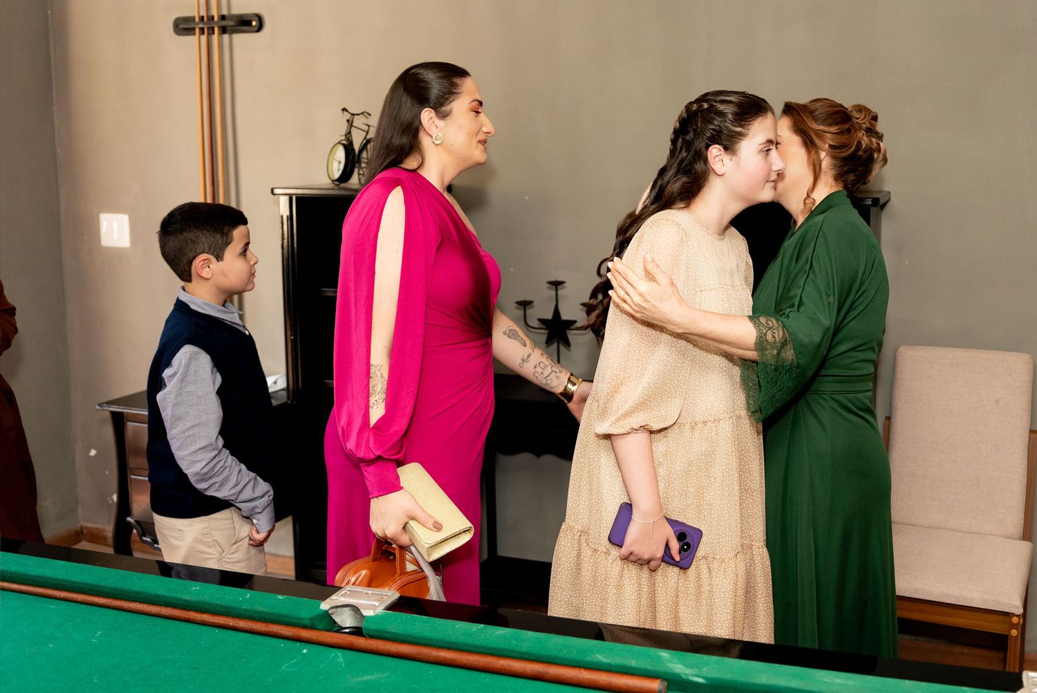 Women in elegant dresses socializing near a pool table, with a young boy standing nearby at a formal gathering.