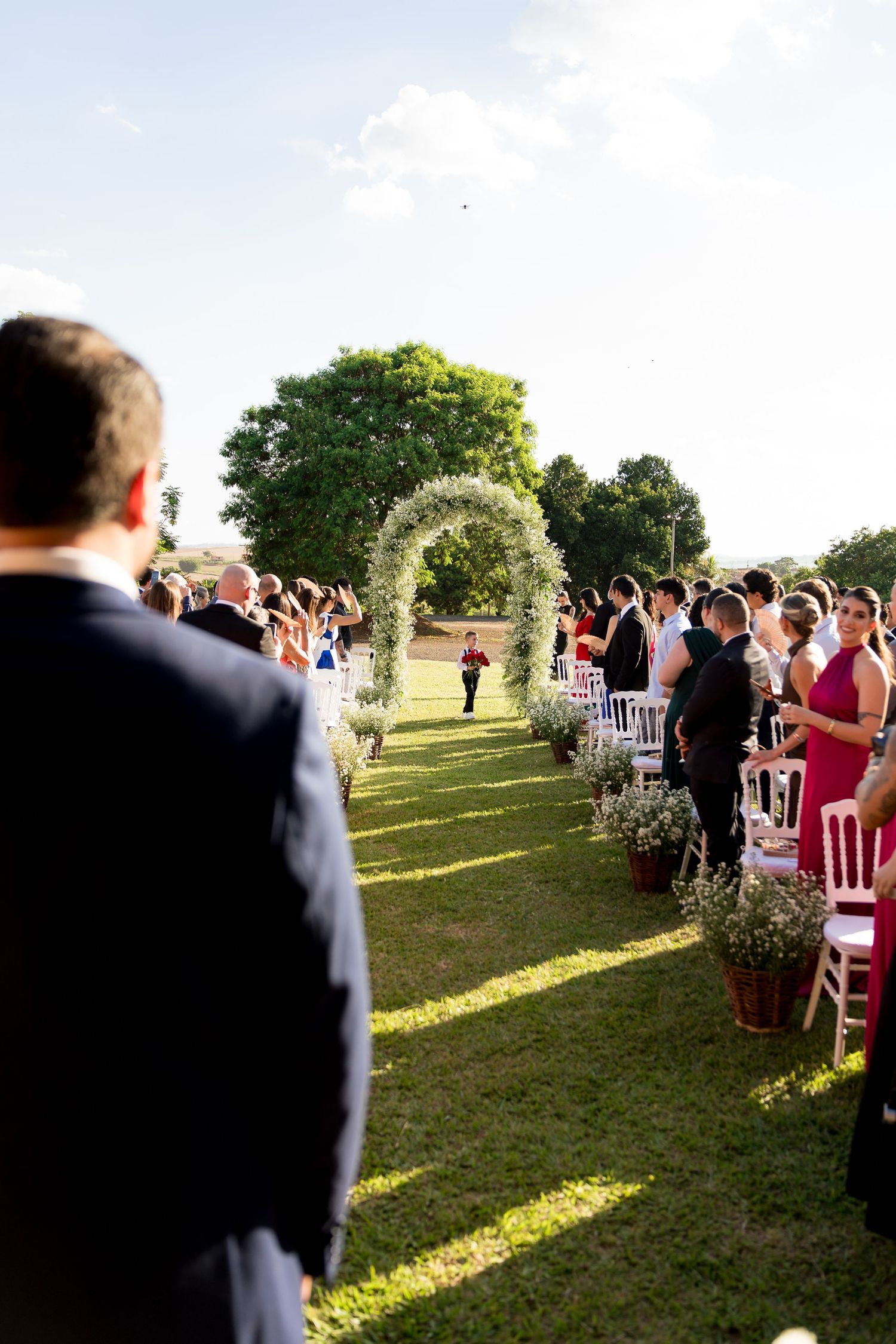 Outdoor wedding ceremony on green lawn with floral arch, white chairs, and guests in formal attire under a clear sky.