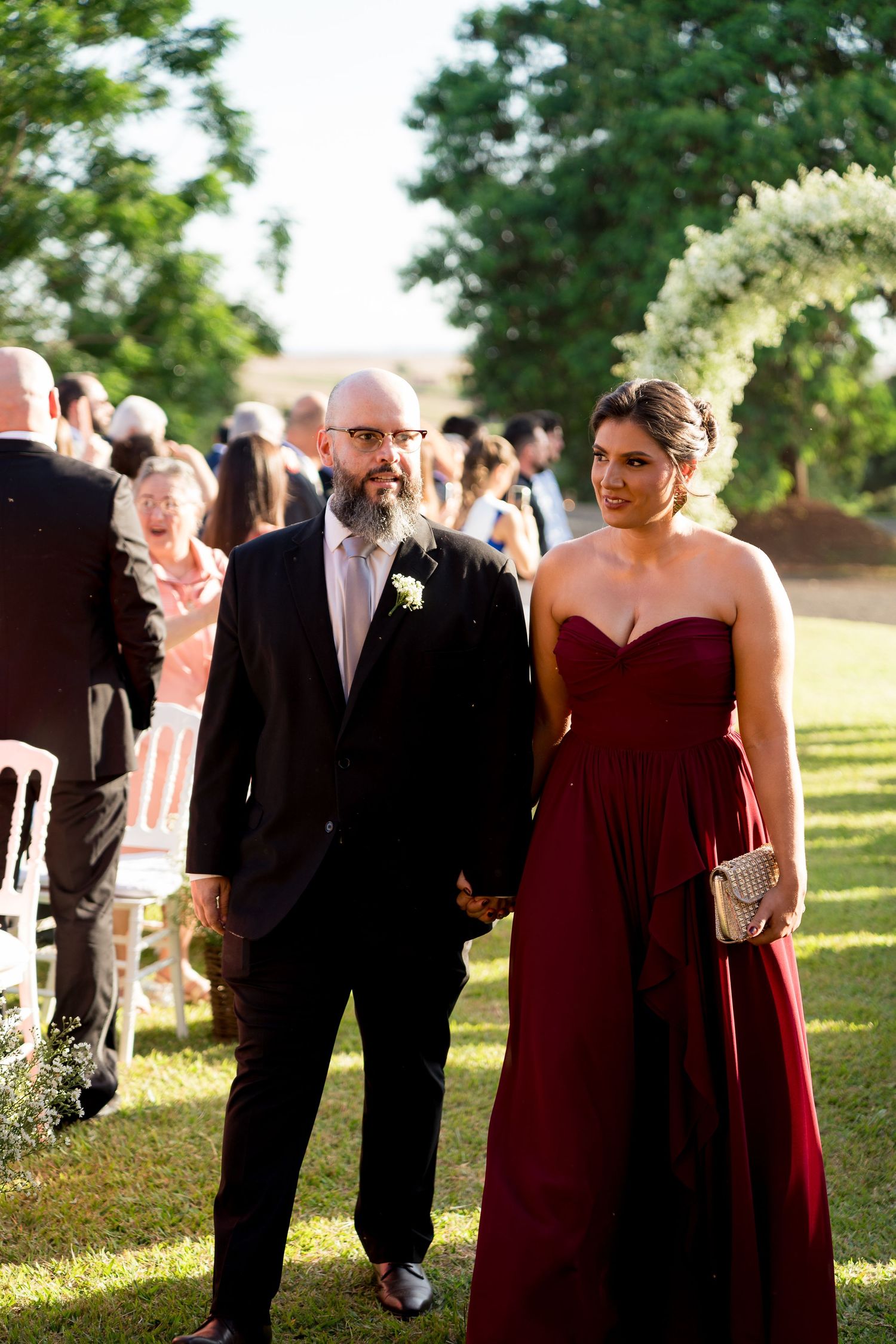 A bald man with a beard in a black suit escorts a woman in a strapless burgundy gown at an outdoor wedding.