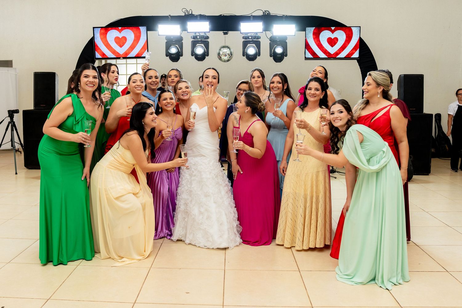 Bride poses with bridesmaids in colorful gowns at wedding reception with heart-shaped backdrop.