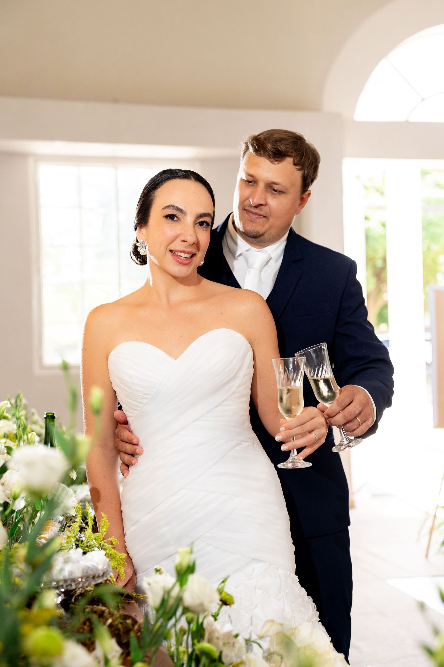 Bride in white strapless gown and groom in navy suit holding champagne glasses at elegant wedding reception.