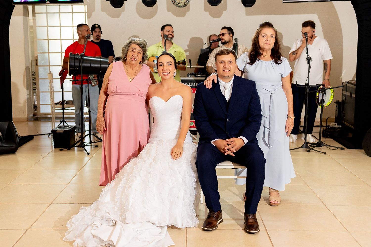 Bride and groom pose with family members at wedding reception, with live band performing in background.